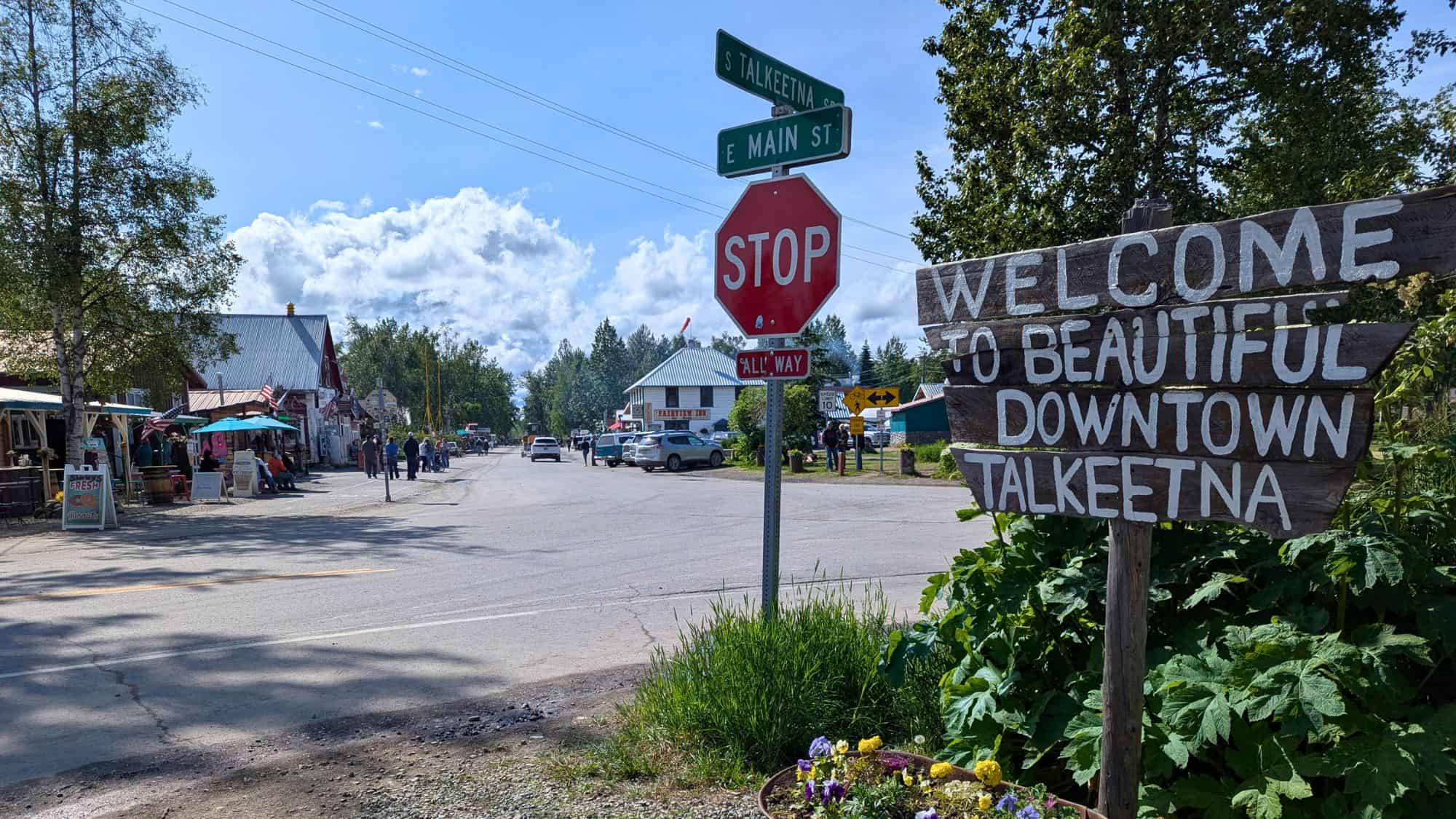 A quaint Alaskan main street scene features a hand-painted wooden sign reading “Welcome to Beautiful Downtown Talkeetna,” a red stop sign, and locals walking past small shops and outdoor seating under flags and umbrellas.