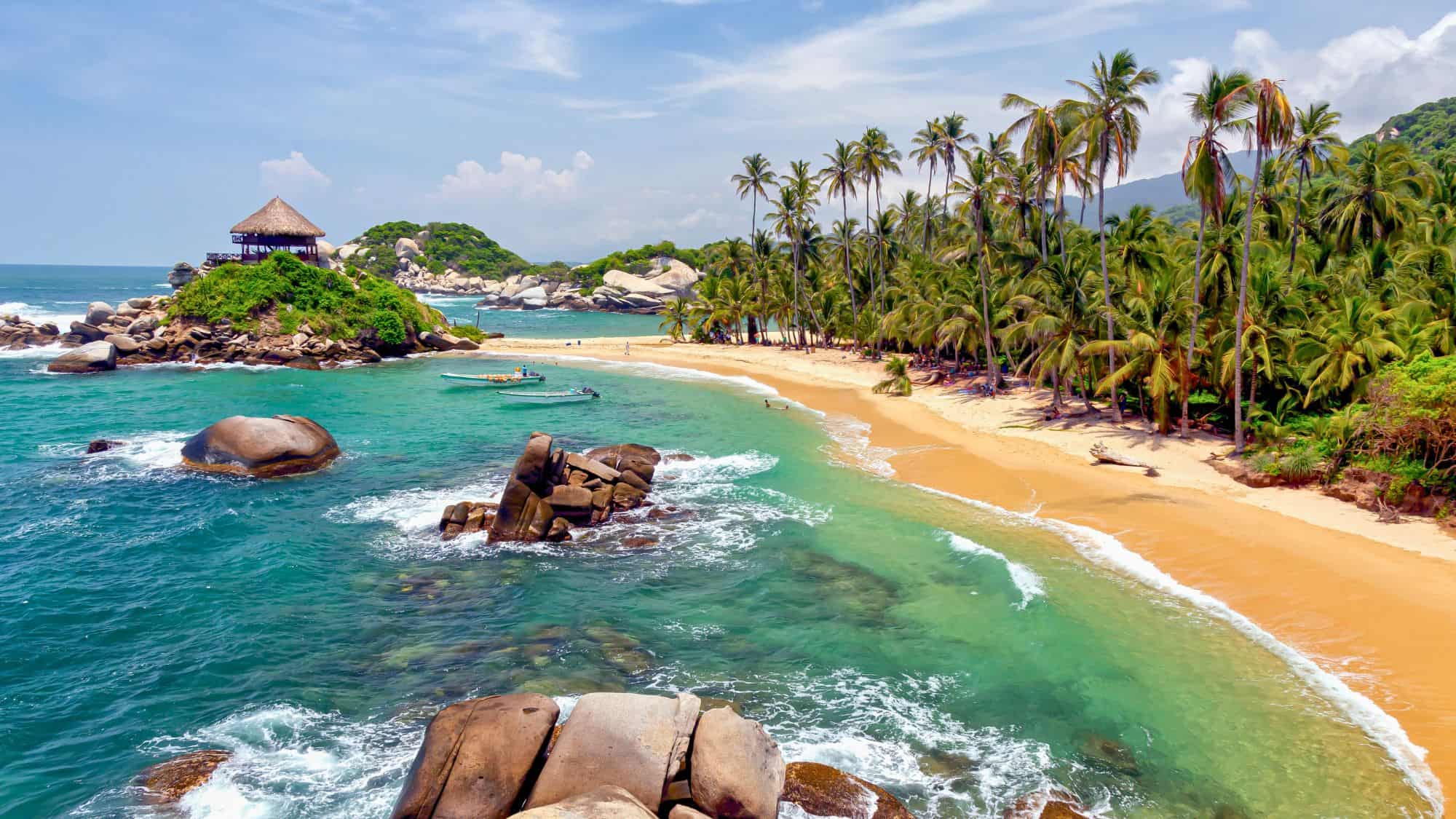 Golden sand curves around a turquoise bay lined with tall palm trees, rocky outcrops, and a thatched-roof hut perched on a jungle-covered hill.