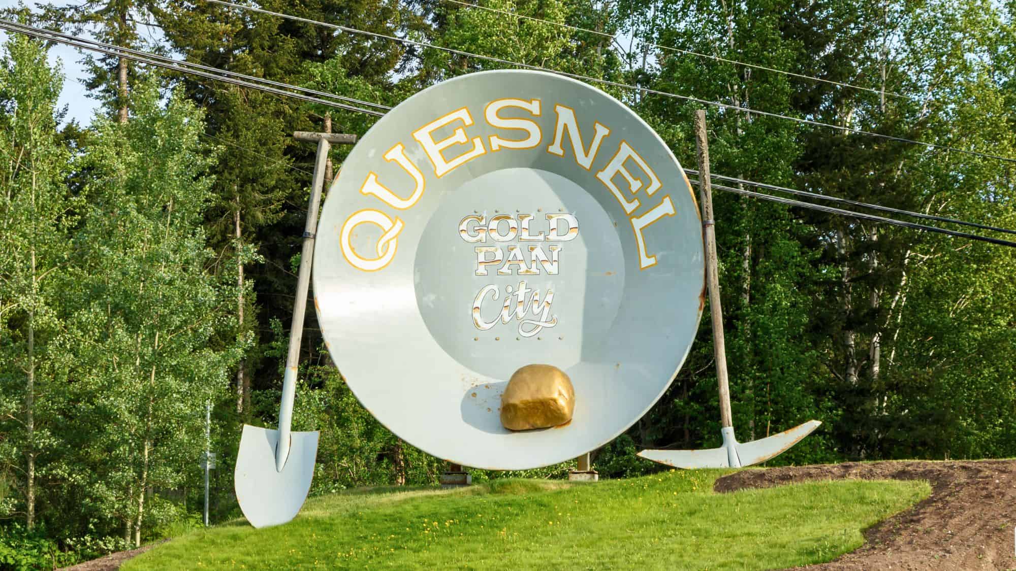 A giant metal gold pan sculpture labeled "Quesnel Gold Pan City" sits on a grassy slope, flanked by two oversized shovels and backed by dense forest—celebrating the town’s gold rush heritage.