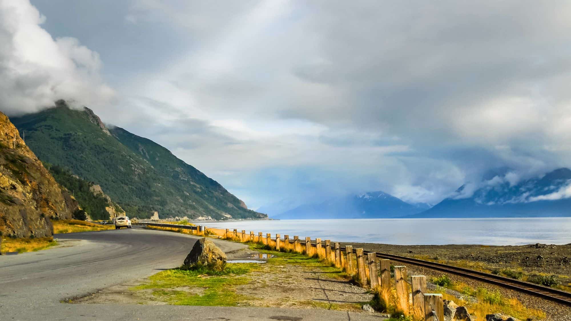 A scenic coastal road curves alongside train tracks and Turnagain Arm, with mist-covered mountain ranges and low-hanging clouds enhancing the moody, atmospheric view.