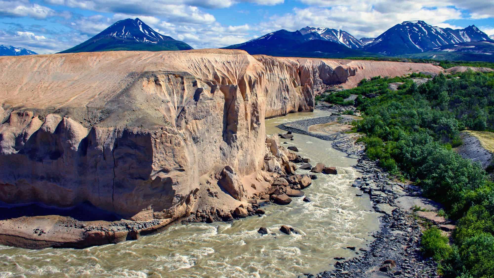 A powerful, muddy river snakes through a deep canyon with eroded tan cliffs, framed by vibrant greenery and snow-speckled mountains in the distance.