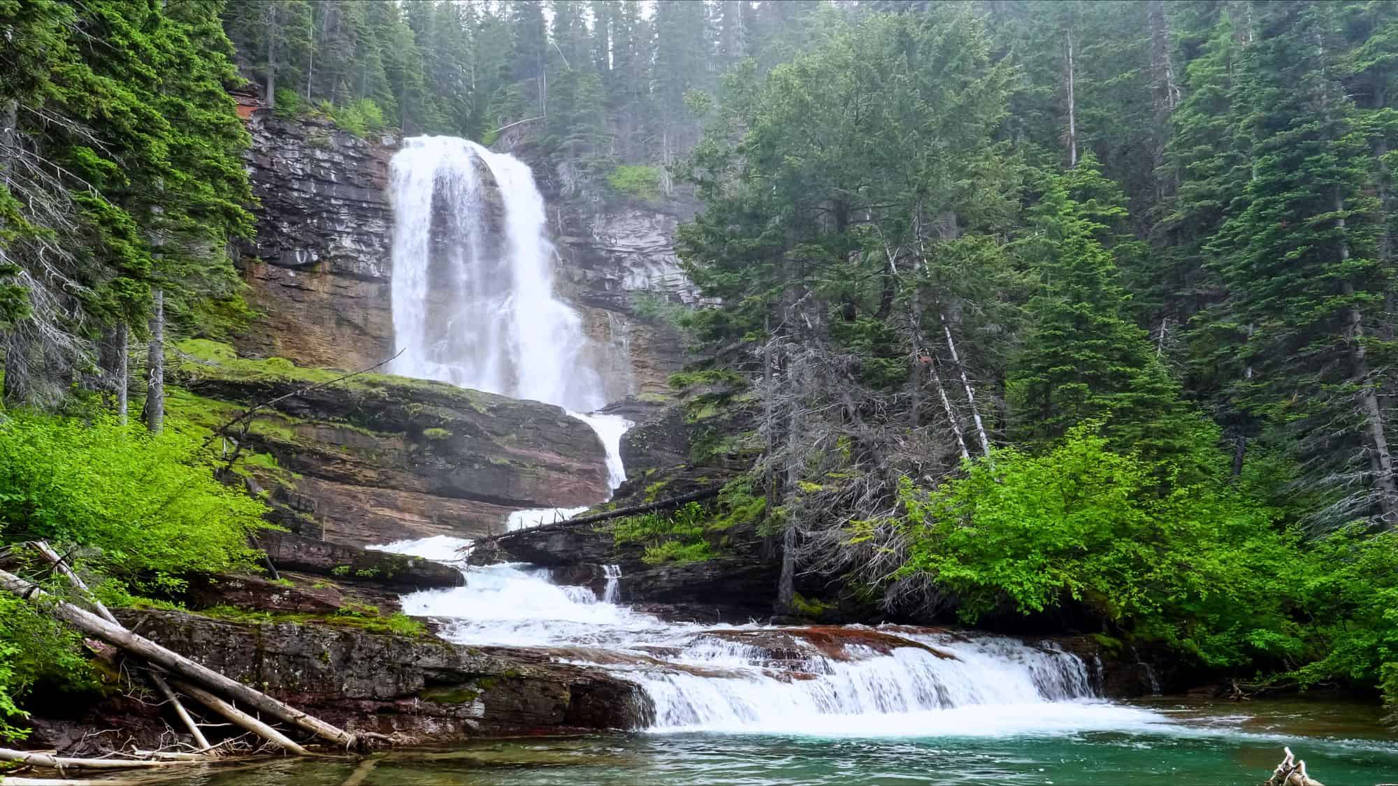 A dramatic multi-tiered waterfall rushes down a red rock cliff into a clear pool, nestled within a thick evergreen forest.