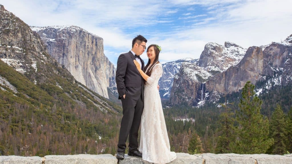 A bride and groom smile at each other while standing before Yosemite's snow-dusted cliffs and pine forest, with El Capitan and Cathedral Rocks visible in the background.