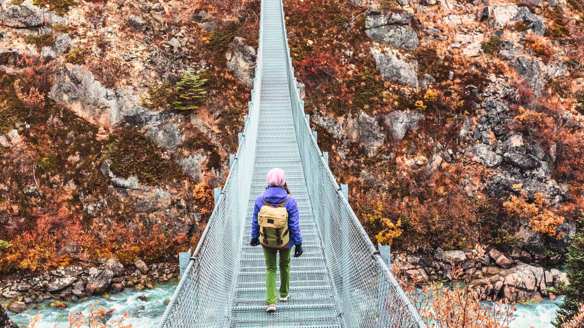 A lone hiker wearing a purple jacket and backpack crosses a narrow metal suspension bridge over a rocky river gorge, surrounded by autumn-colored trees and steep rock walls in the Yukon wilderness.