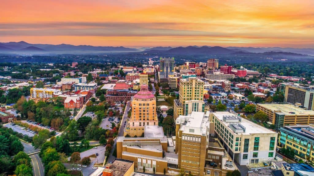 A vibrant aerial view of downtown Asheville, North Carolina, with the city’s eclectic architecture framed by colorful sunset skies and the distant Blue Ridge Mountains.