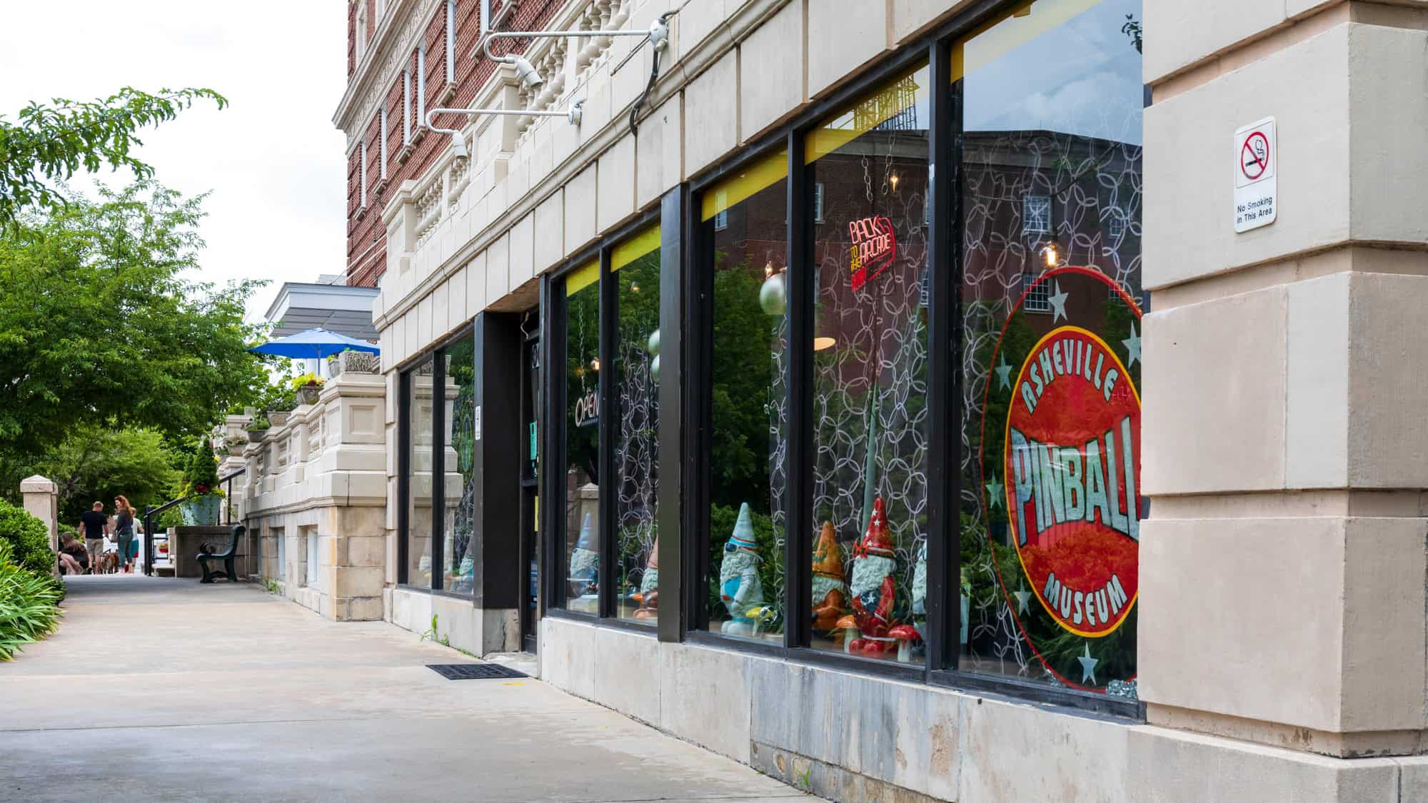 A sidewalk view of the Asheville Pinball Museum's window display, featuring colorful gnome figures and a red circular sign with bold white text, set in a historic downtown building.