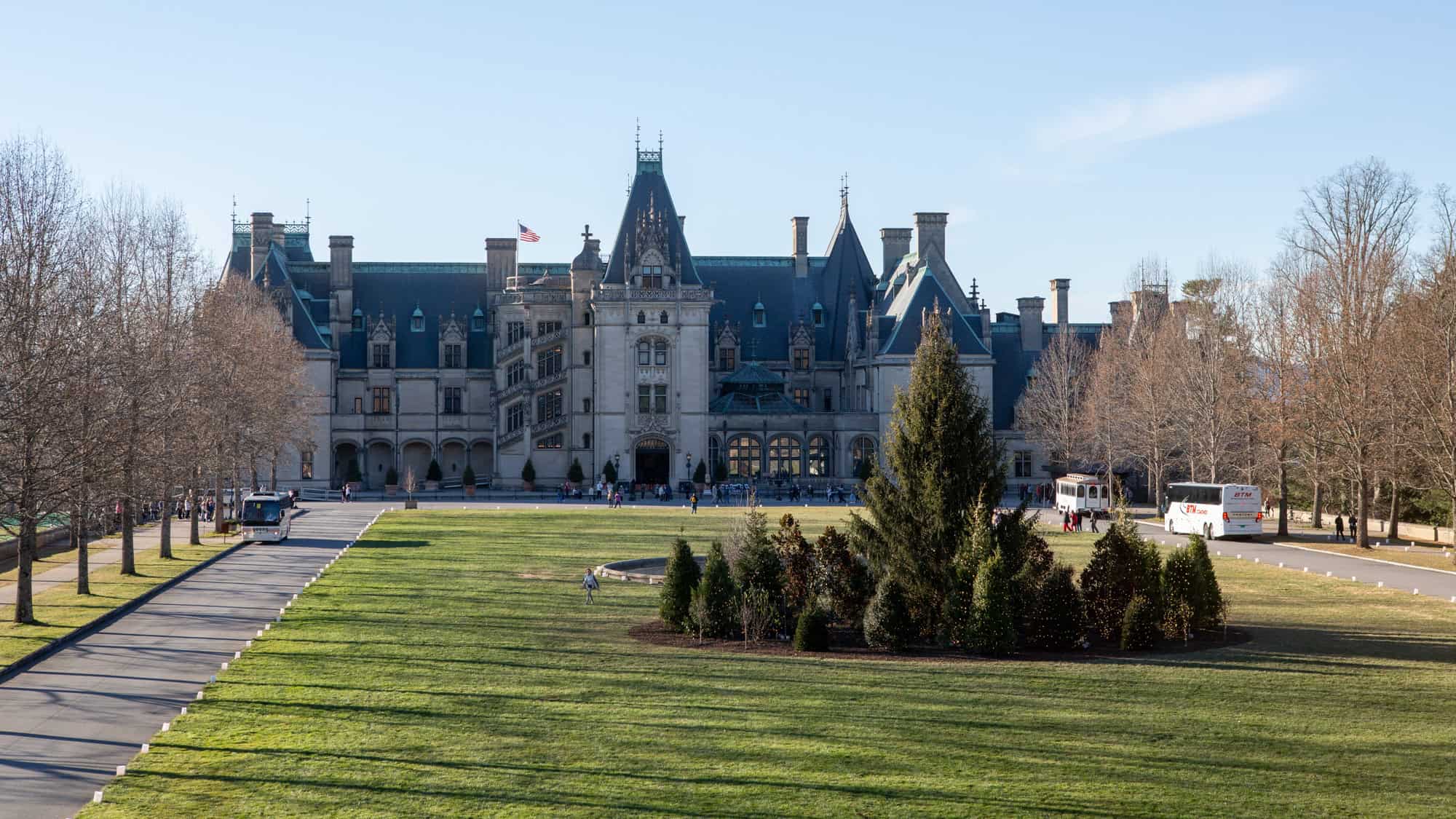 A grand chateau-style mansion with a steeply pitched roof and ornate architecture stands at the end of a manicured lawn, with visitors, buses, and a U.S. flag visible near the entrance.