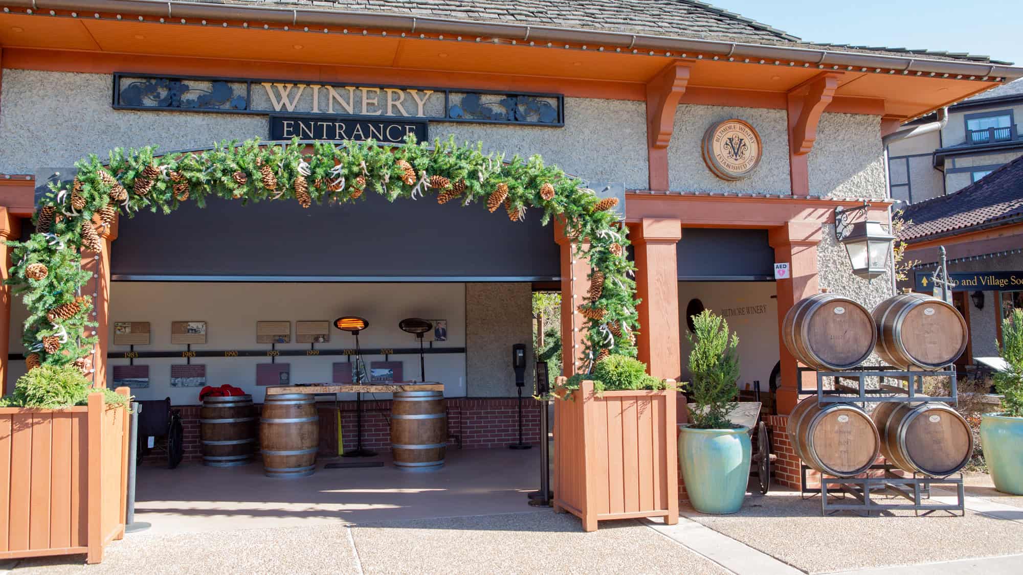 The entrance to Biltmore Winery is decorated with festive pinecone garland and wooden barrels, with a timeline display visible inside the open-air tasting area.