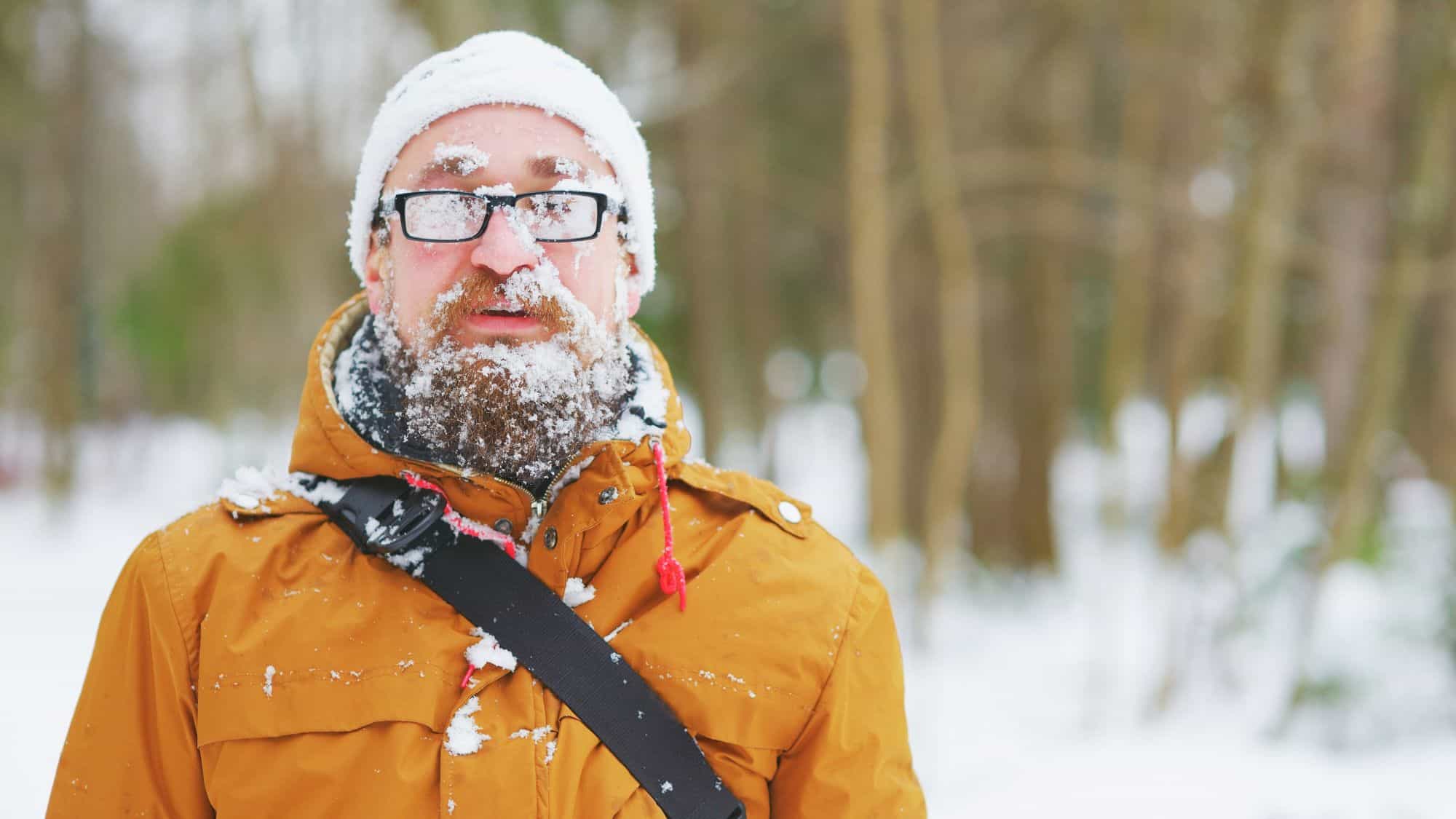 A man bundled in a mustard parka and white hat is covered in snow and ice crystals, with glasses fogged over in a wintry forest setting.