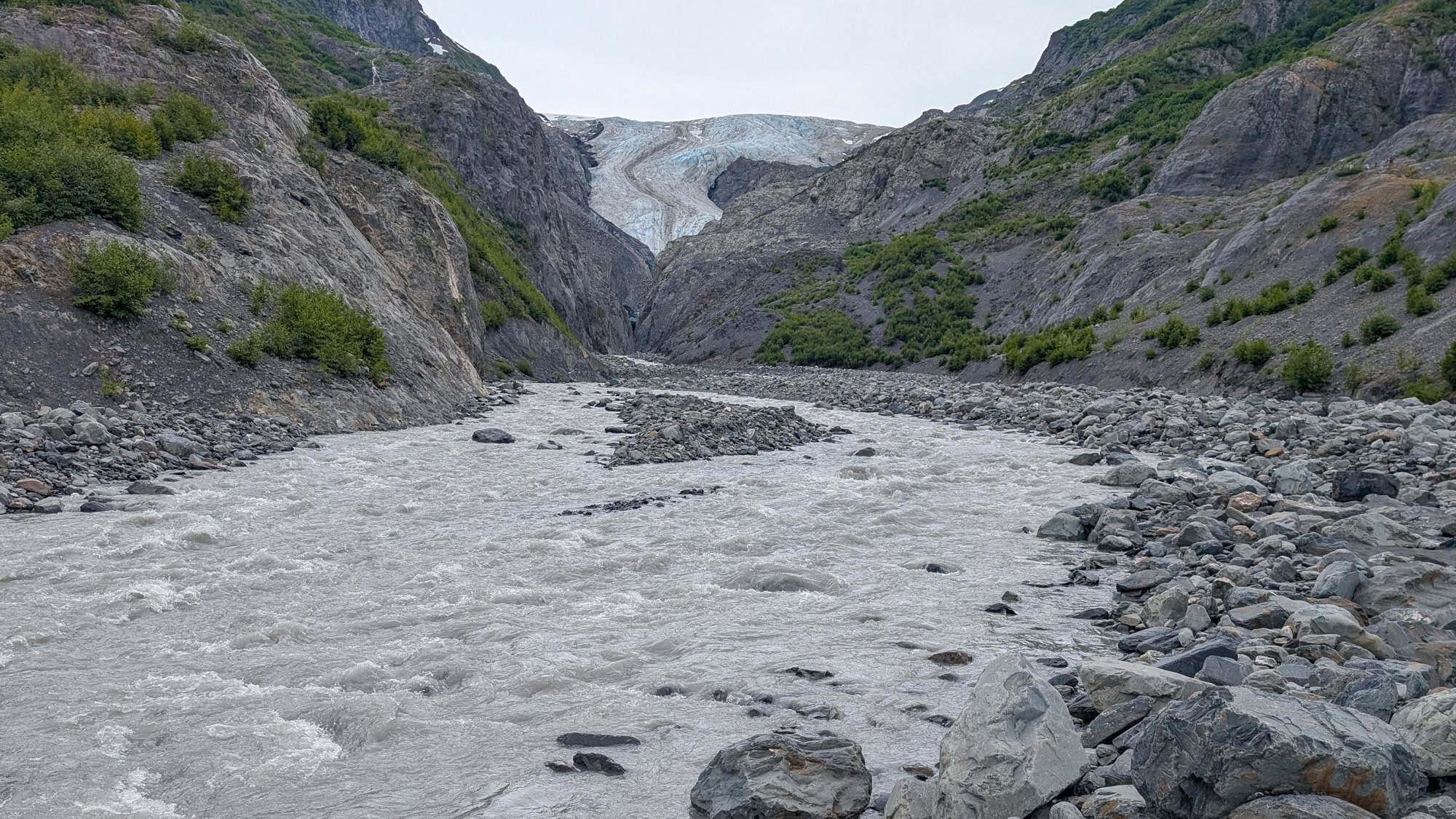 A silty, fast-moving glacial river cuts through rocky terrain, fed by the distant Exit Glacier nestled between rugged cliffs.