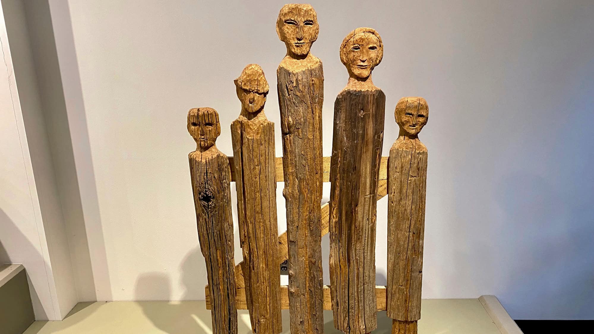 A rustic wooden sculpture featuring six vertical, carved figures with stylized faces, created from rough timber, displayed against a plain white museum wall.
