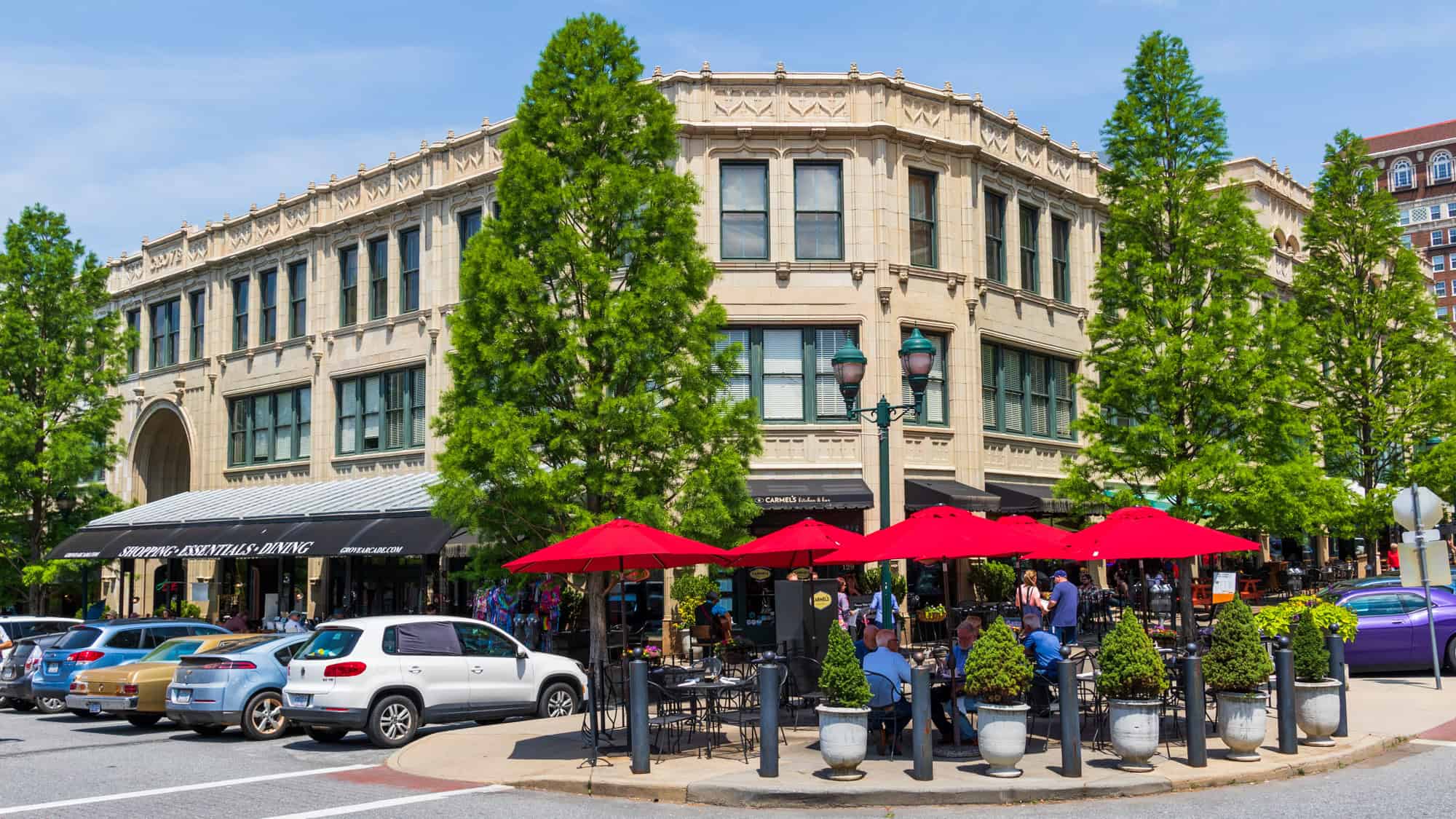 Street-level view of the historic Grove Arcade building in downtown Asheville, with red umbrellas shading outdoor diners and tall green trees lining the sidewalk.