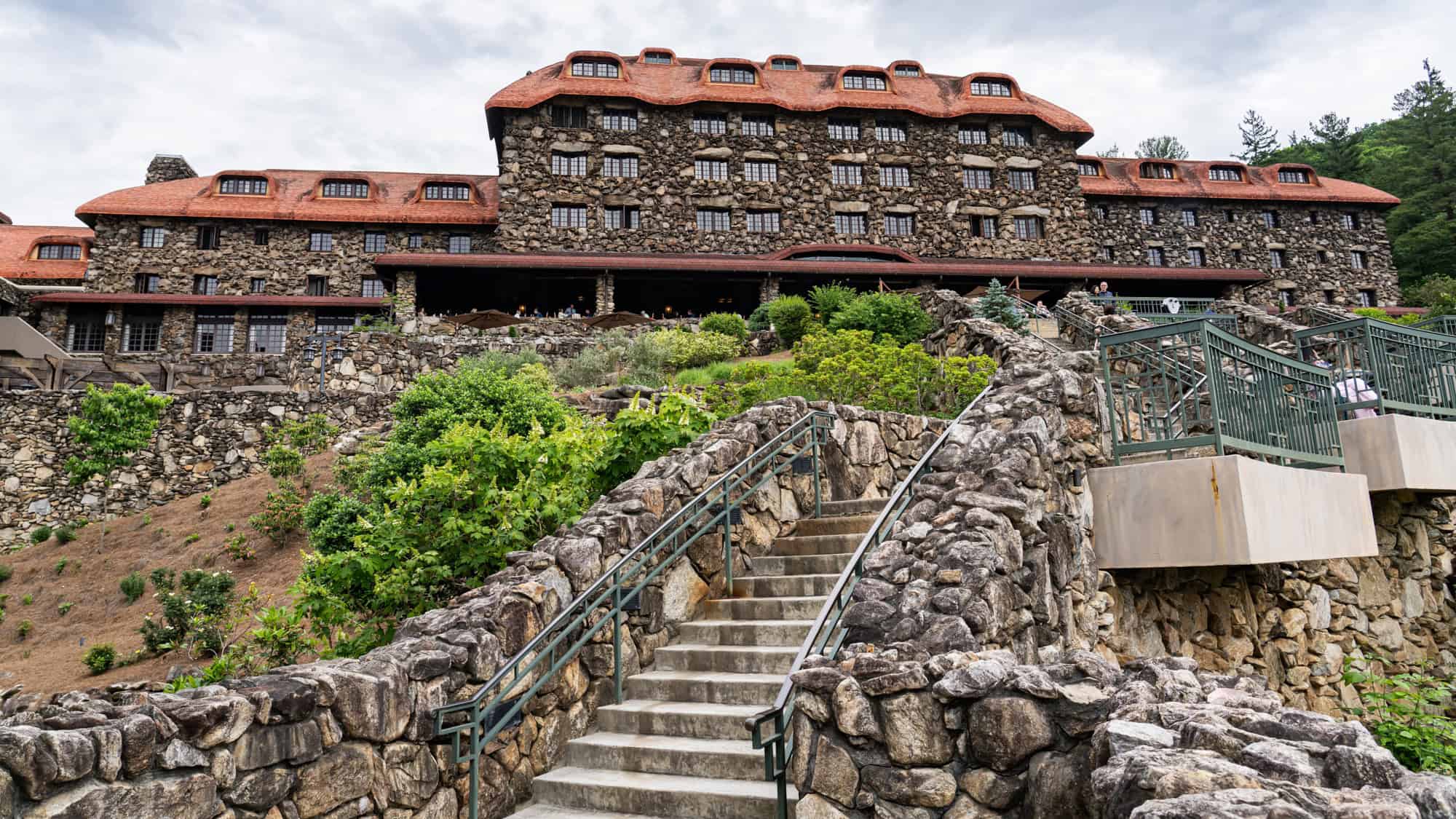 The stone facade and terracotta roof of the historic Grove Park Inn rise behind terraced stone stairways and railings, surrounded by greenery and set against a cloudy sky.