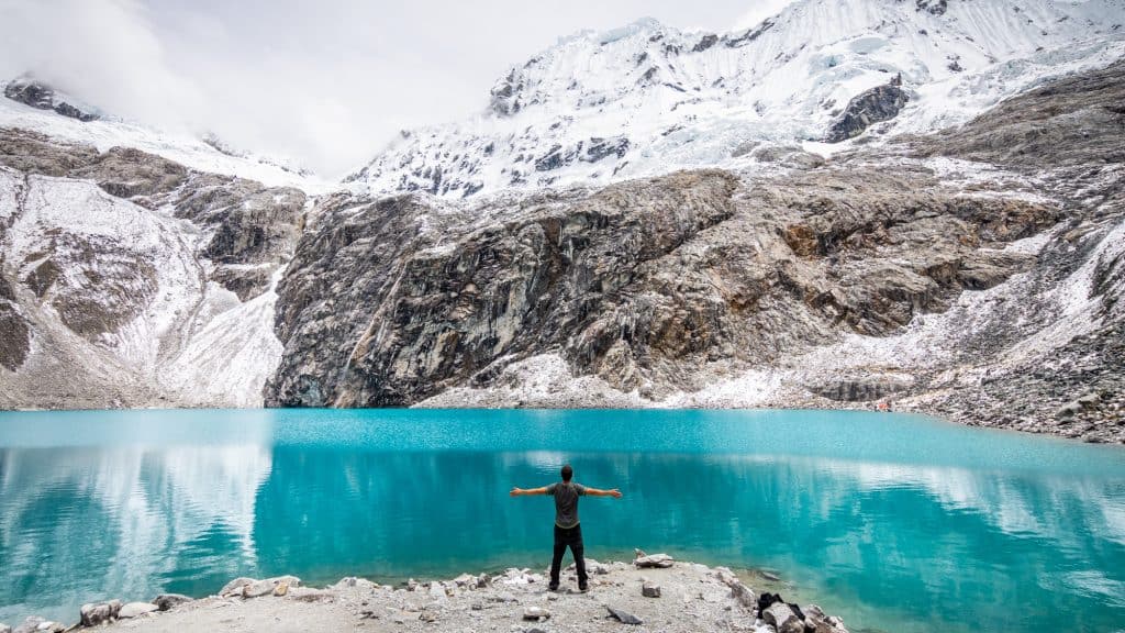 A person stands with arms outstretched at the edge of Laguna 69 in Peru, facing a vivid turquoise glacial lake backed by snow-covered rocky peaks and misty clouds in the Cordillera Blanca range.