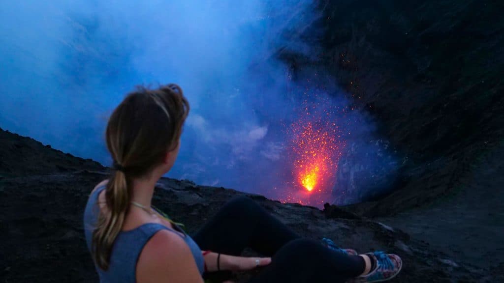 A woman sits near the edge of a dark volcanic crater, watching molten lava glow and spark inside the depths, partially shrouded in volcanic steam.