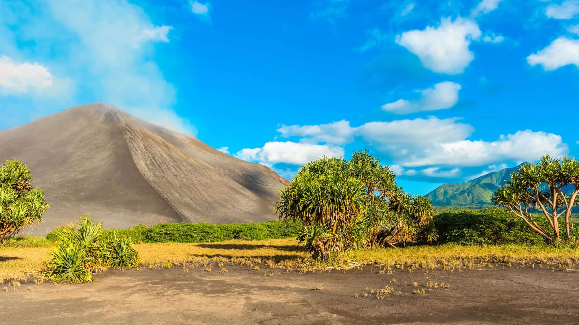 The steep, ash-gray cone of Mount Yasur rises behind bright green tropical trees and shrubs under a sunny, blue sky with scattered clouds.