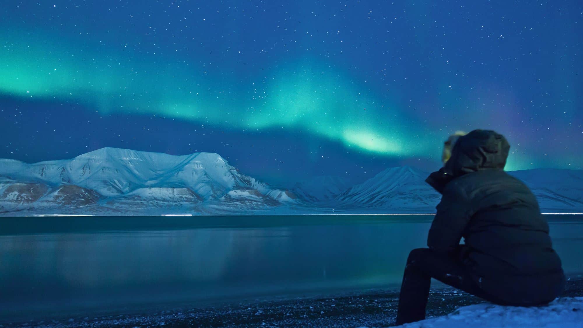 A person in a parka sits by a frozen fjord watching vivid green northern lights ripple above snow-covered peaks under a clear star-filled sky.