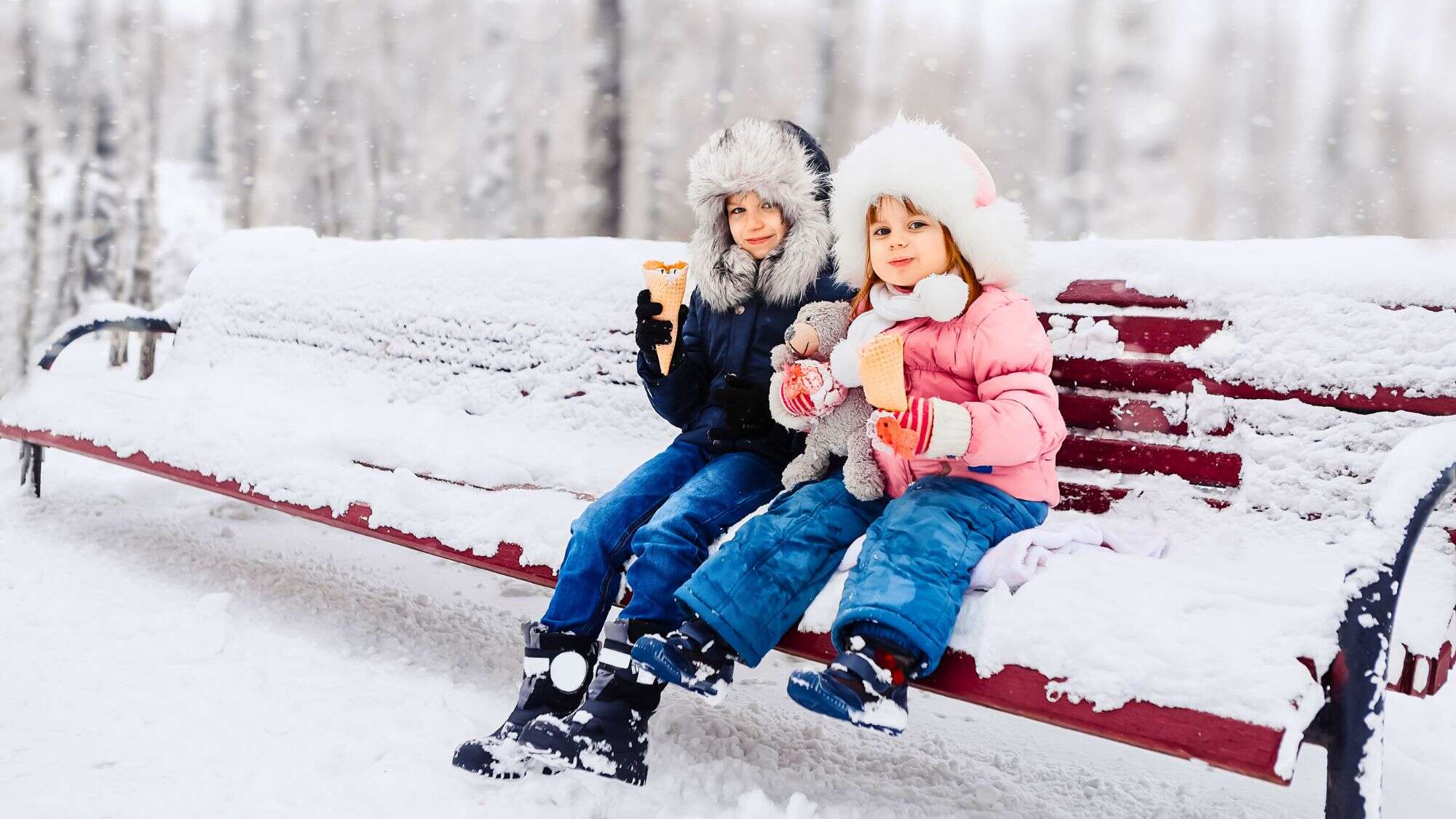 Two kids in puffy coats and furry hats sit on a snow-covered bench in a snowy park, happily eating ice cream cones while one holds a stuffed animal.