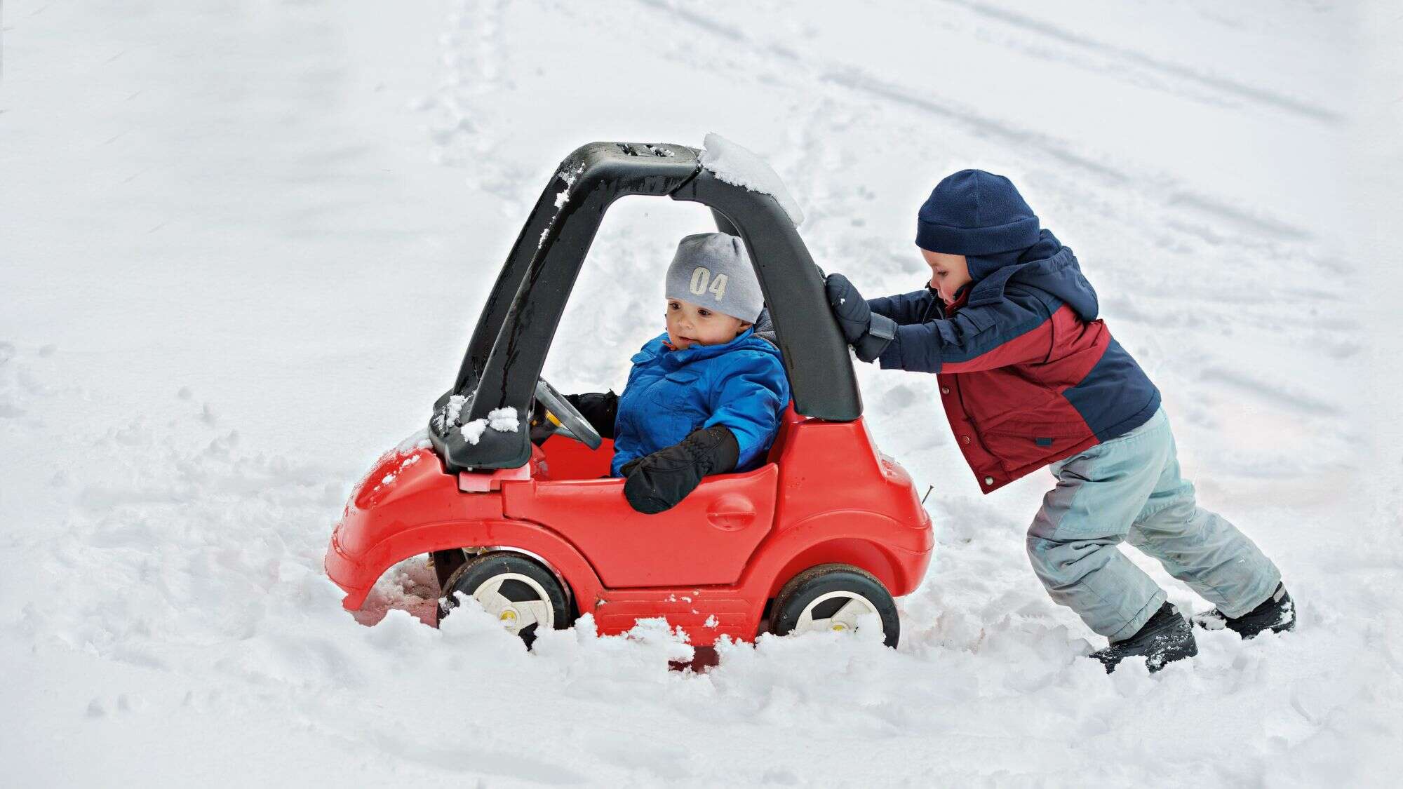 A bundled-up child pushes a red toy car through fresh snow while another child rides inside, both dressed in winter coats and hats.