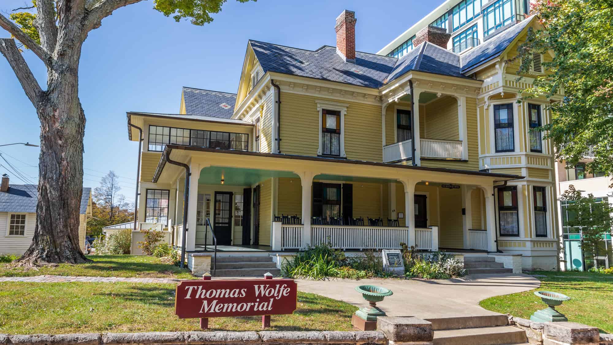 A preserved yellow Victorian house with a wraparound porch and dark trim, identified by a red sign in front as the Thomas Wolfe Memorial, sits on a manicured lawn under clear blue skies.
