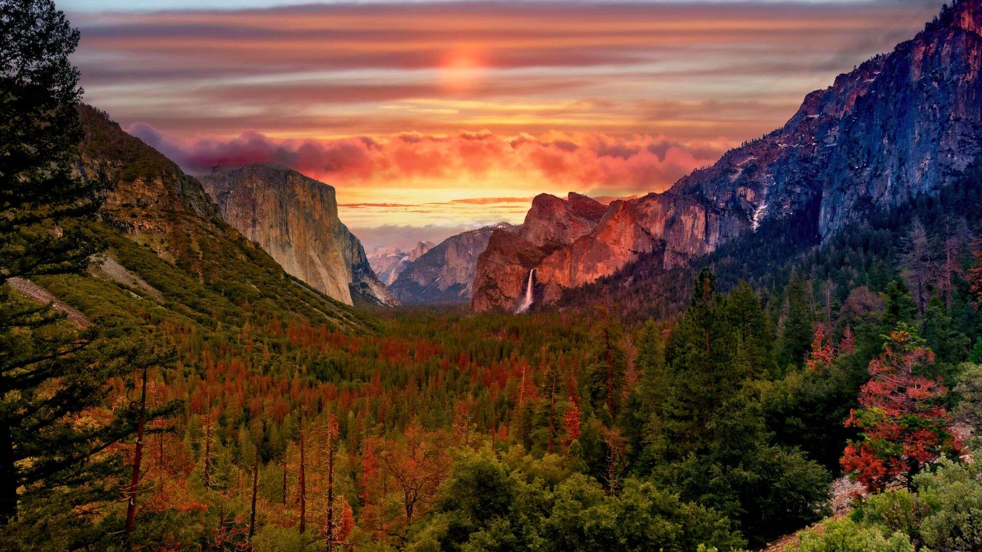 A vivid sunset bathes Yosemite Valley in golden and magenta tones, highlighting El Capitan, Bridalveil Fall, and distant peaks under a sky streaked with colorful clouds.