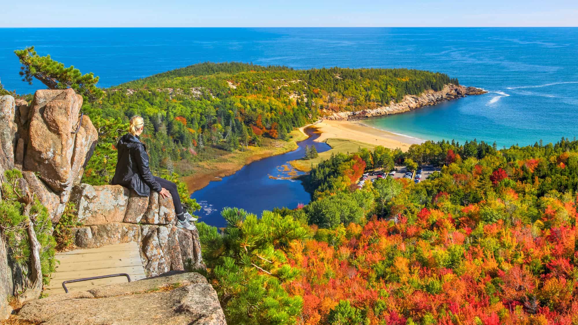 A woman sits on a rocky cliff admiring a sweeping view of autumn-colored forests, a sandy beach, and turquoise ocean waters.