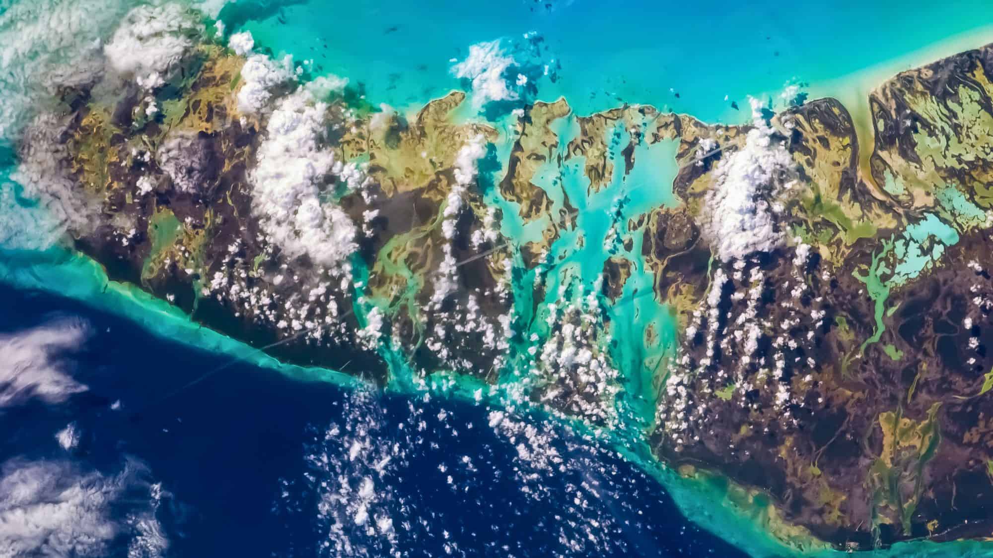 A bird’s-eye view shows a sprawling reef system with vivid blue shallows, dark land patches, and scattered white clouds.