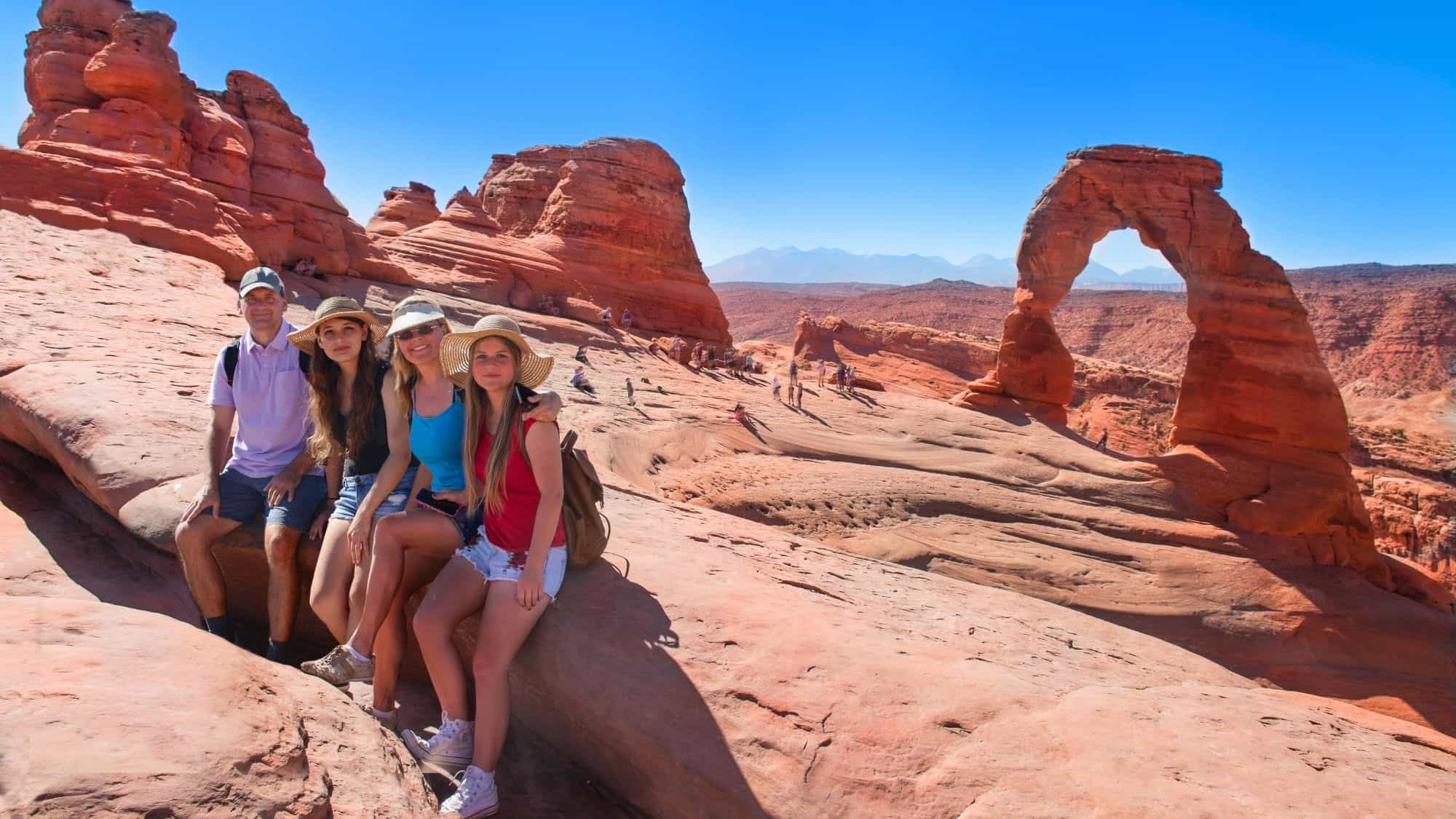 A group of four people sits on sunlit red sandstone near Delicate Arch in Arches National Park, with the iconic rock formation rising against a bright blue sky.