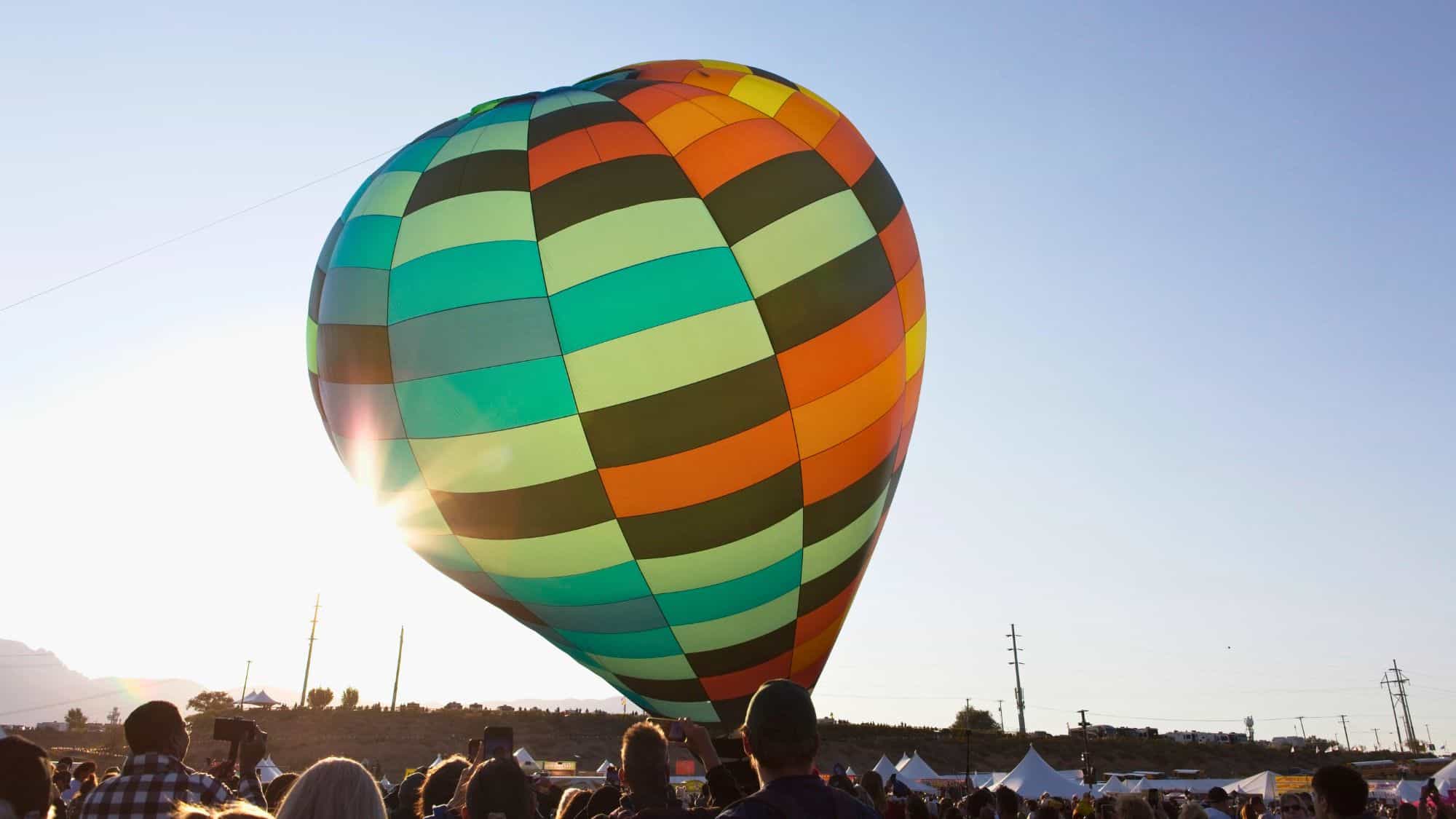 A colorful hot air balloon with green, orange, and yellow panels begins to rise as a crowd watches in the sunlight.