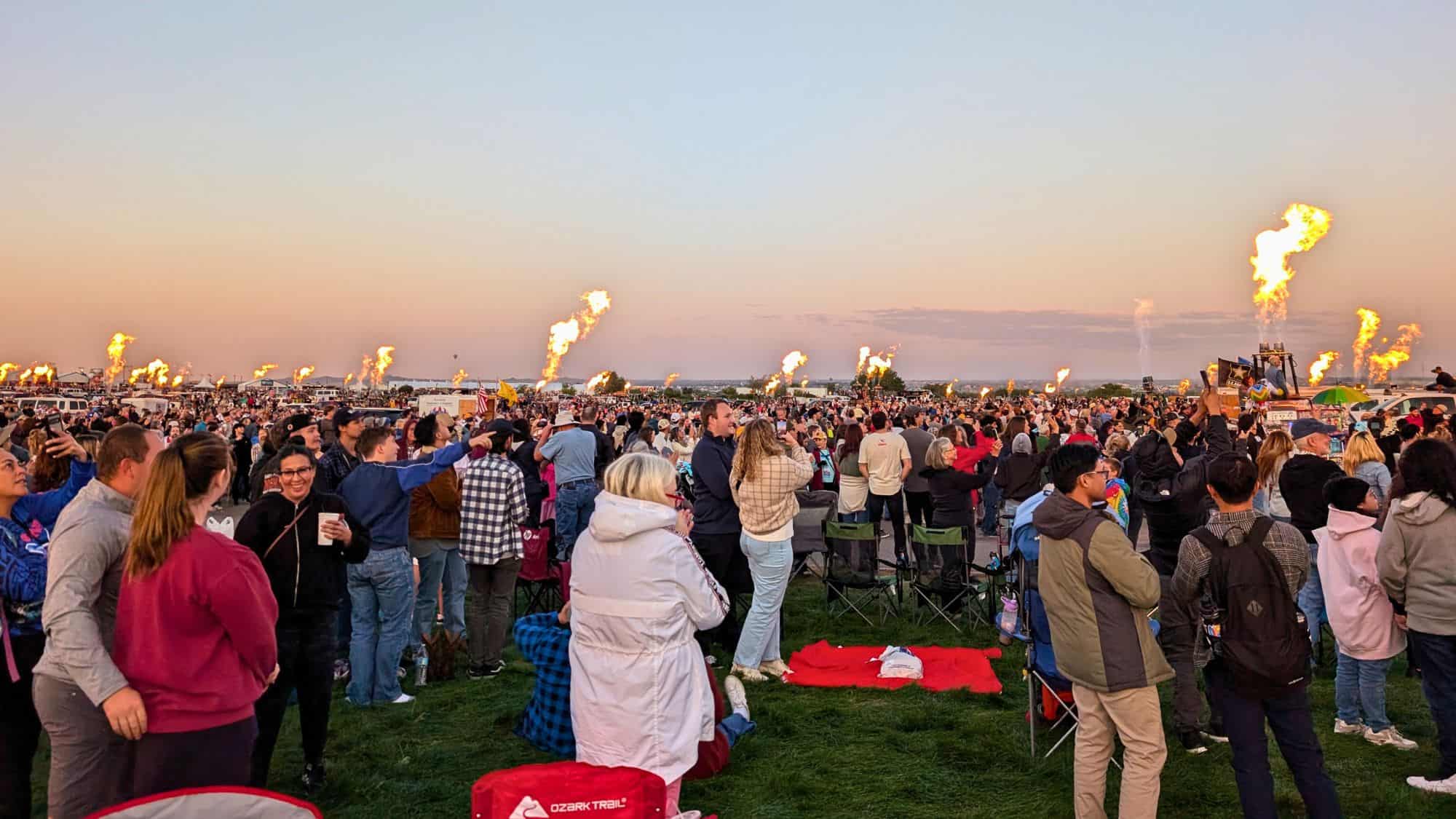 A large crowd gathers at dusk as rows of balloon burners shoot tall flames into the sky.