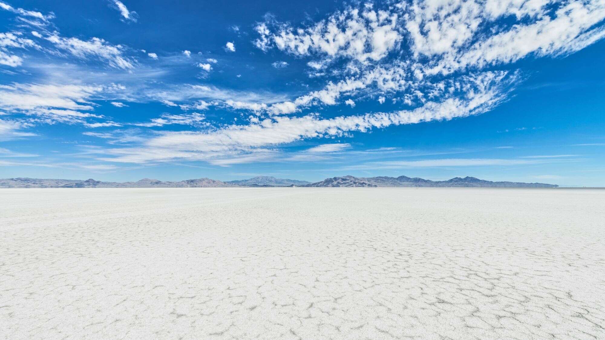 A vast expanse of white salt flats stretches toward distant mountains under a bright blue sky streaked with wispy clouds.