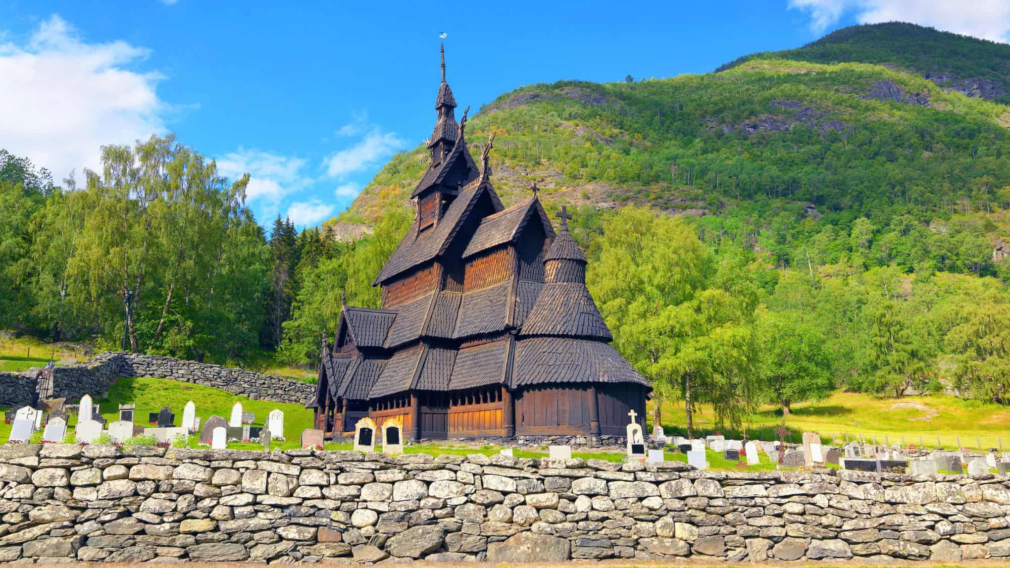 A historic wooden stave church stands in a lush valley, surrounded by gravestones and backed by green hills.