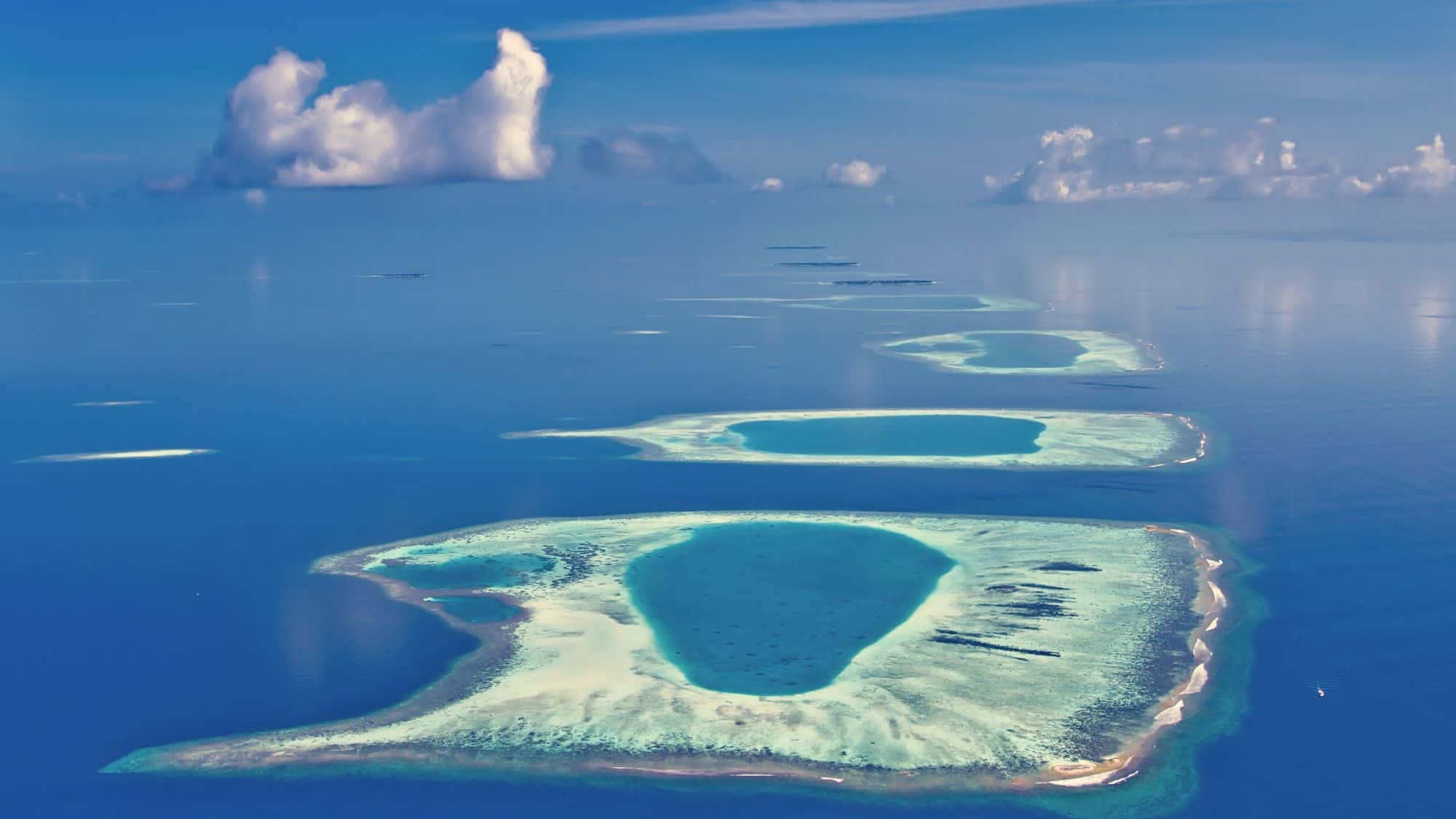 A line of circular coral atolls stretches across the deep blue ocean, fading into the horizon.