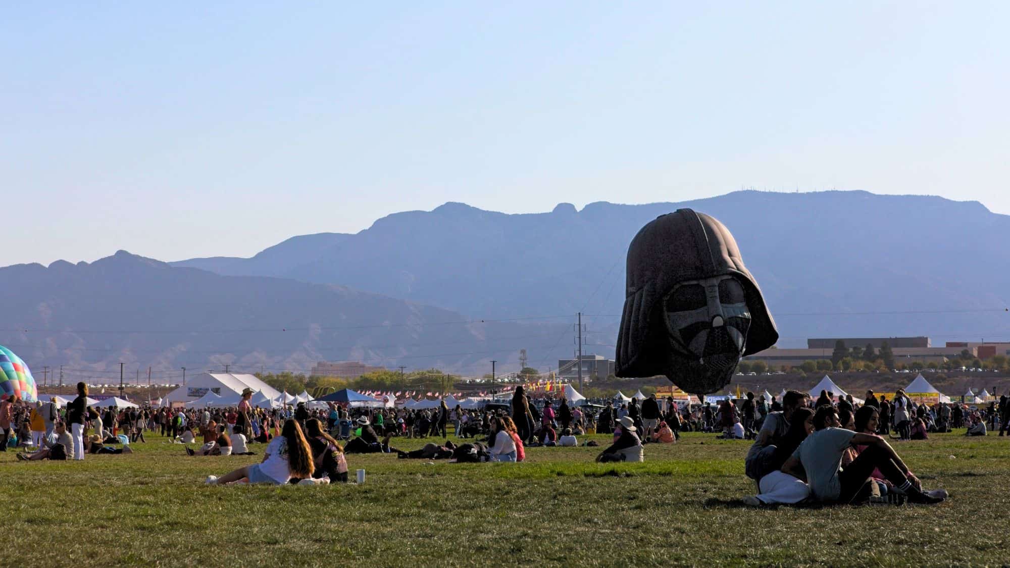 A hot air balloon shaped like Darth Vader’s helmet floats above a grassy field with mountains in the background.