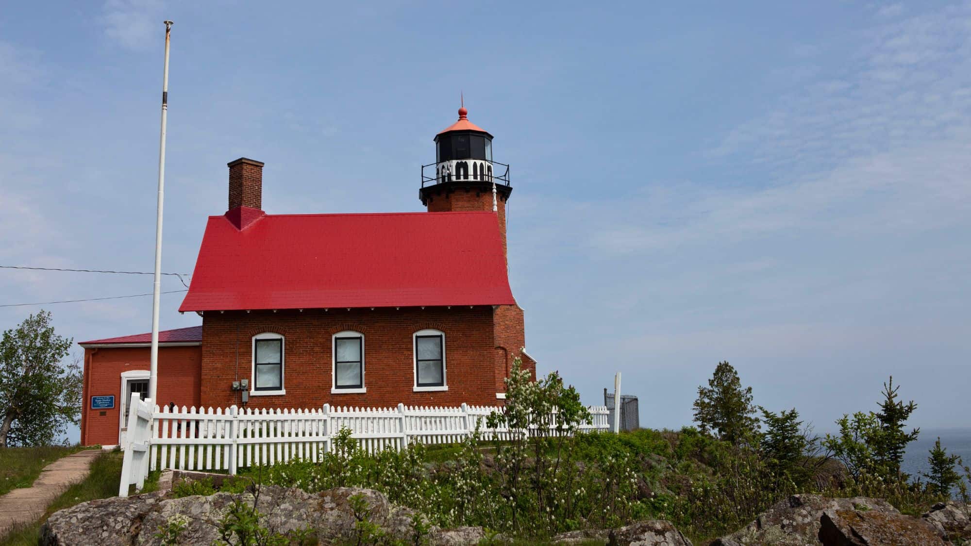 A brick lighthouse with a bright red roof and white picket fence sits on a rocky hilltop, overlooking the water under a partly cloudy sky.