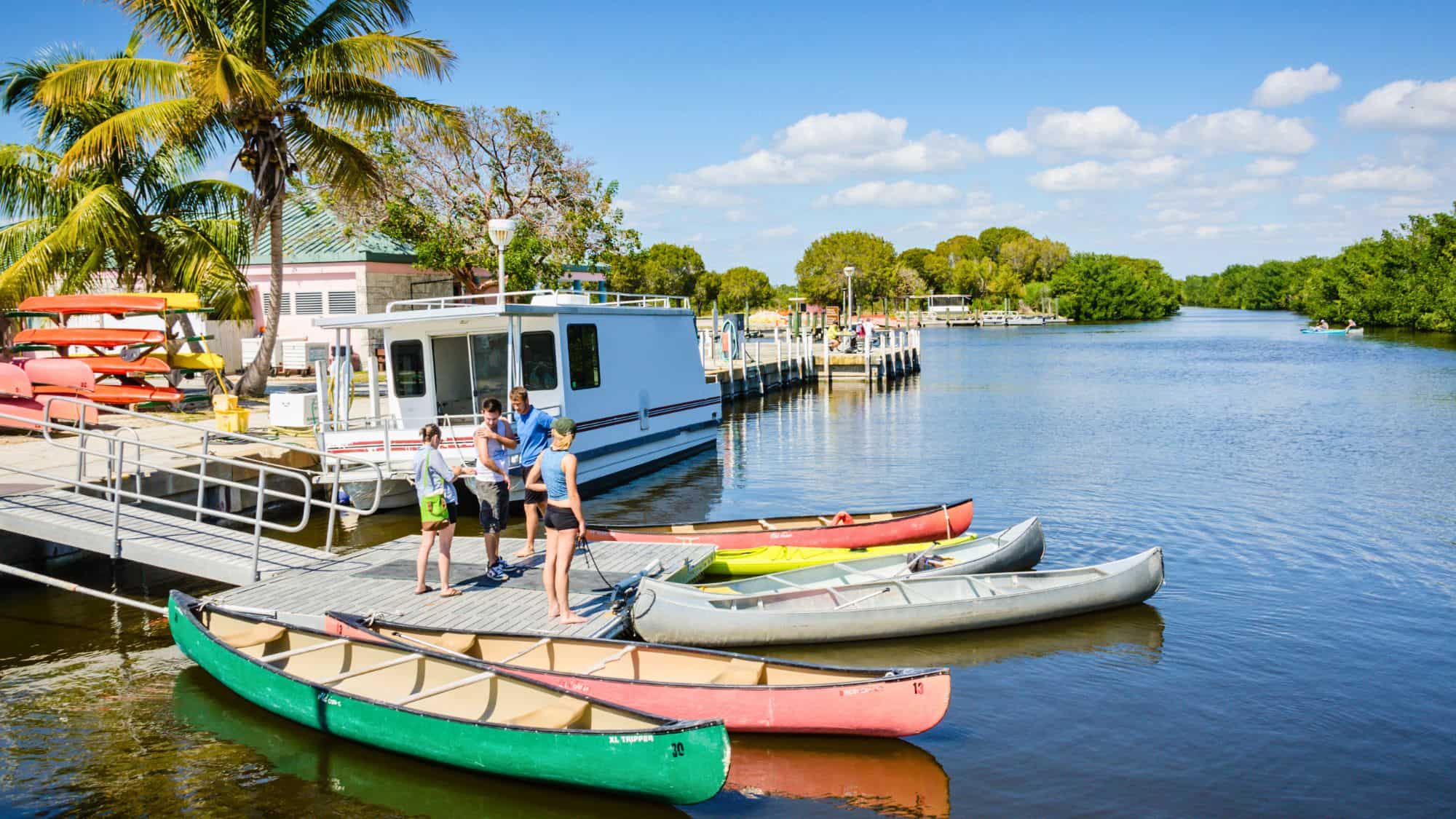 People gather at a dock lined with colorful canoes beside calm water, with palm trees and a houseboat in the background.
