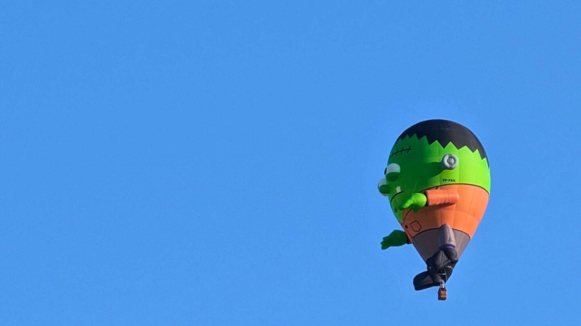 A hot air balloon shaped like a cartoonish green Frankenstein figure drifts against a clear blue sky.