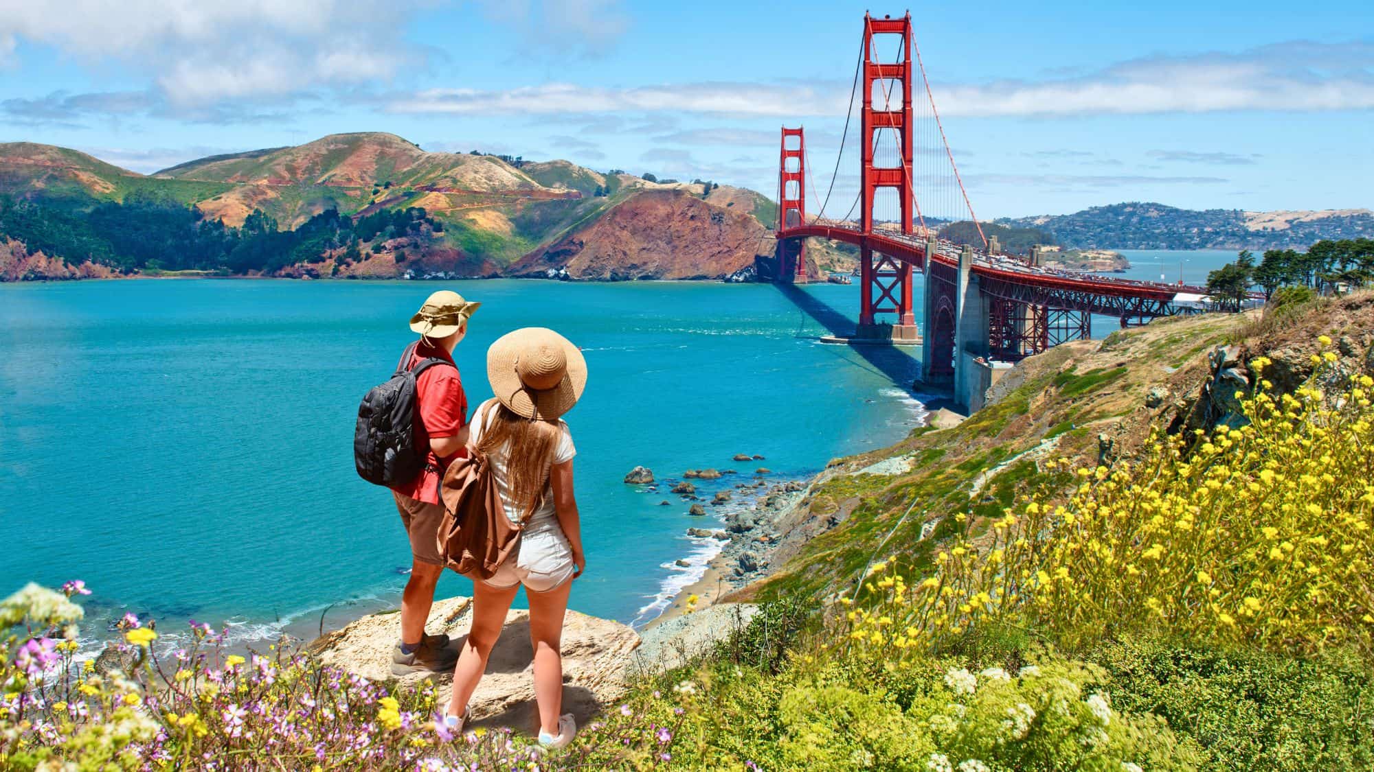 Two hikers stand on a bluff surrounded by wildflowers, overlooking the Golden Gate Bridge and turquoise waters below.