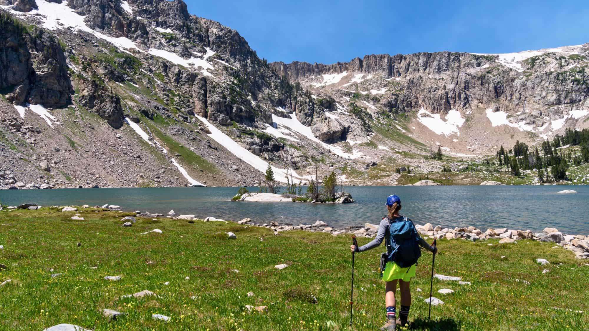 A hiker approaches a tranquil alpine lake with snow patches on surrounding rugged peaks under a bright summer sky.