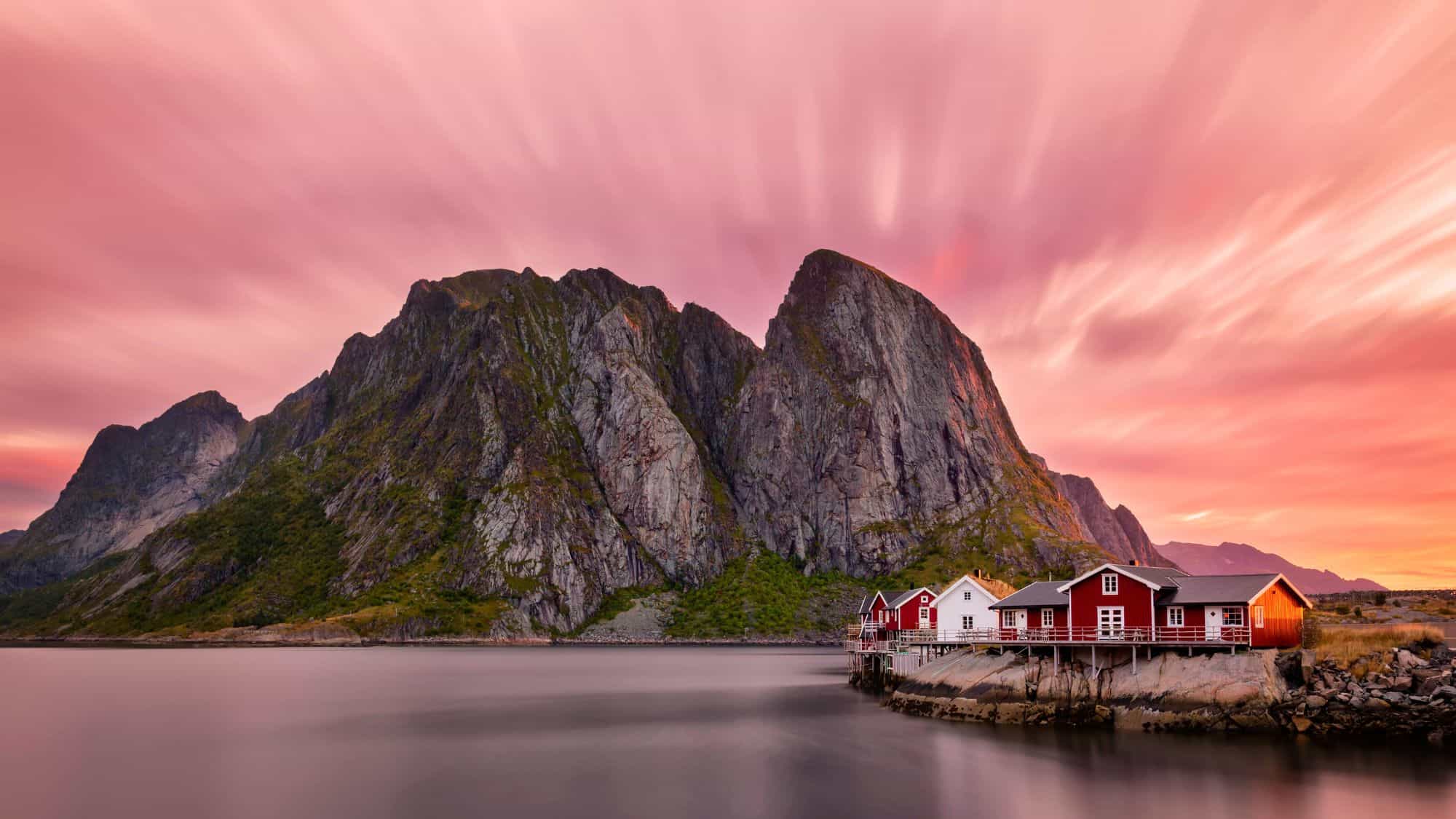 Red fishing cabins sit at the edge of calm water under a pink and orange sky, with towering cliffs behind.