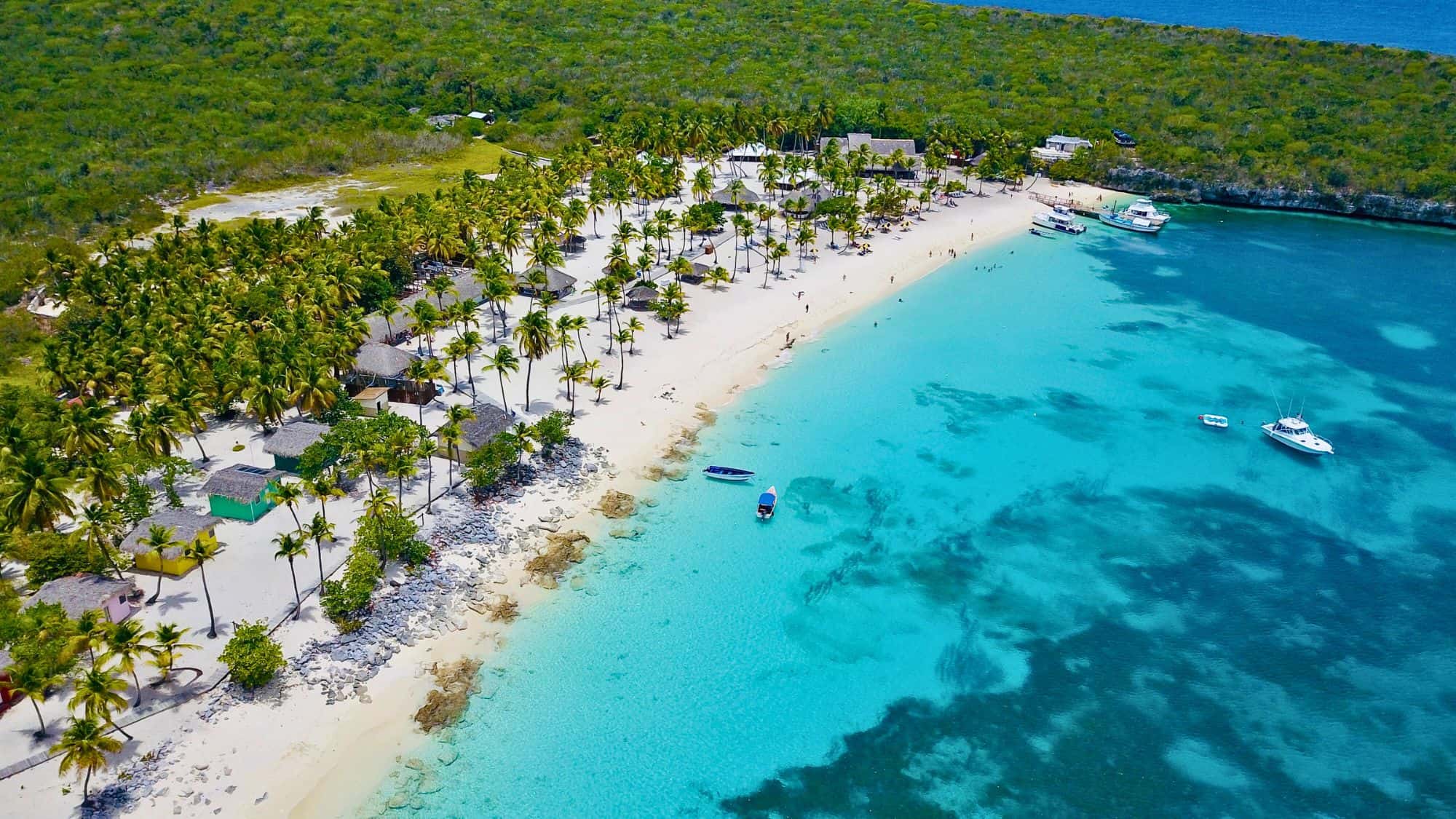 An aerial view of a palm-lined white sand beach with turquoise water, small boats, and scattered thatched huts.