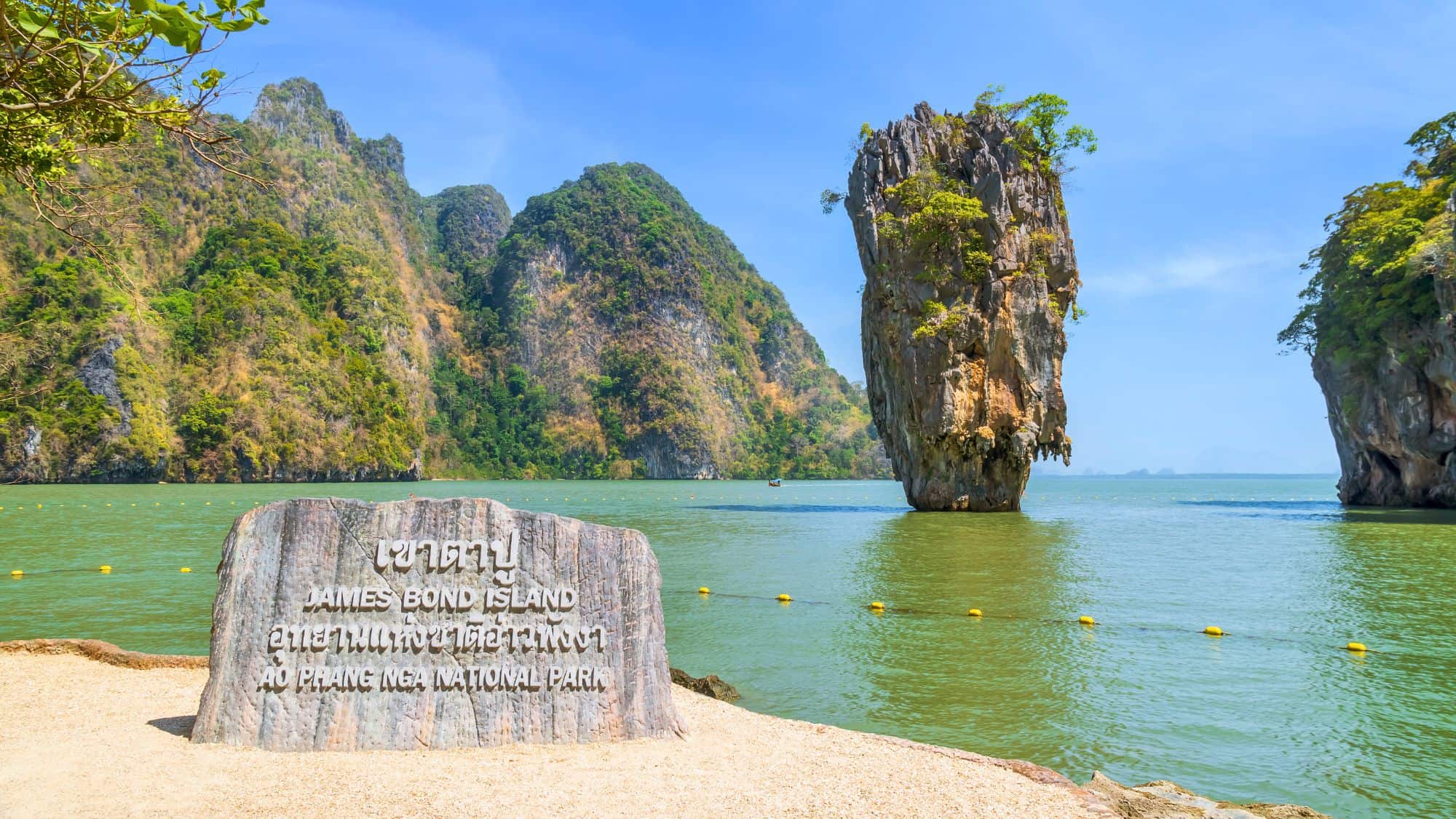A towering limestone karst juts dramatically from emerald-green waters, framed by lush cliffs, with a sign marking its fame as James Bond Island.