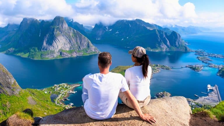 A couple sits on a rocky ledge, admiring the sweeping mountain and fjord views of the Lofoten Islands.