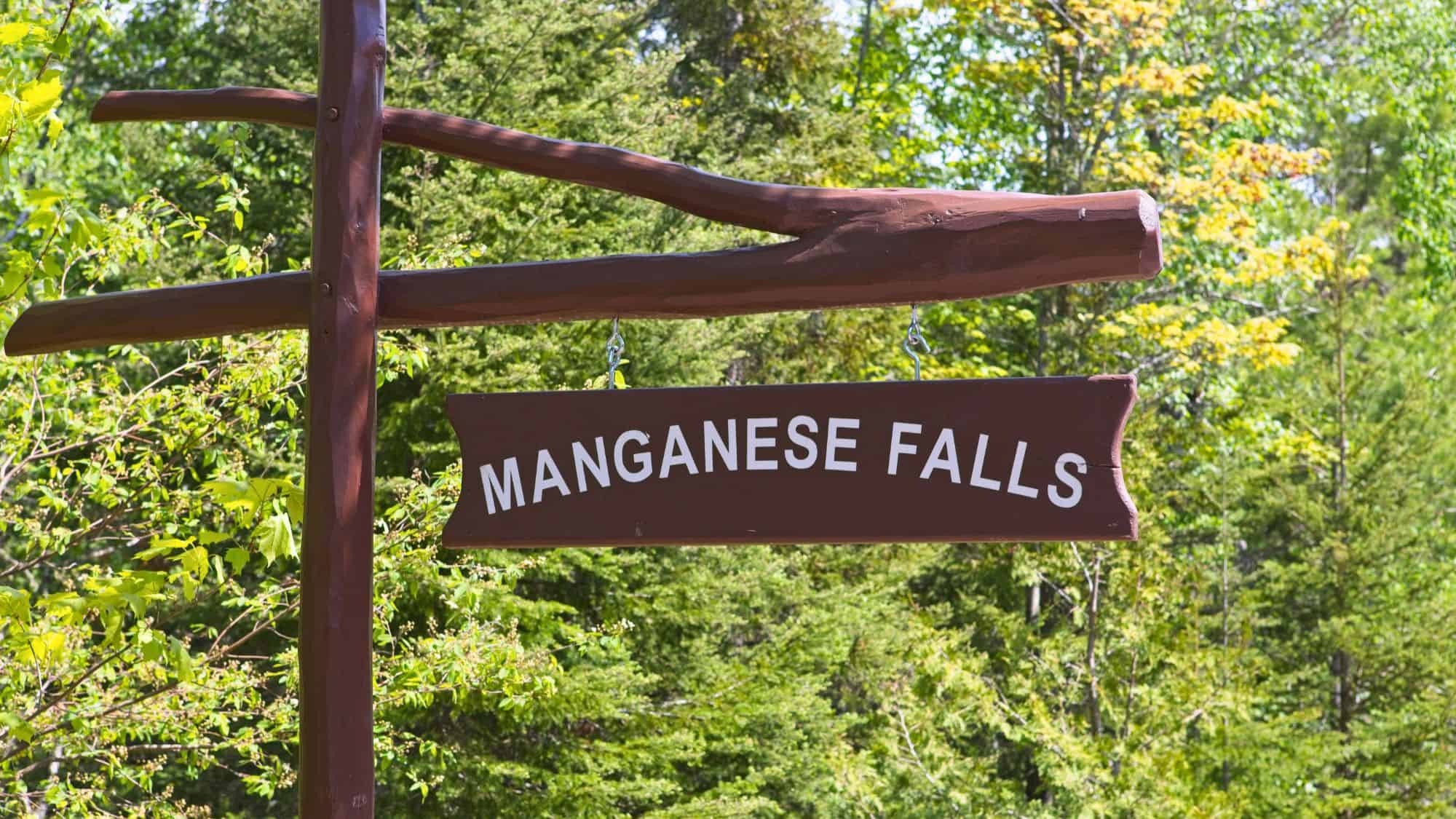 A rustic wooden sign reading “Manganese Falls” hangs from a timber post, surrounded by leafy green trees.
