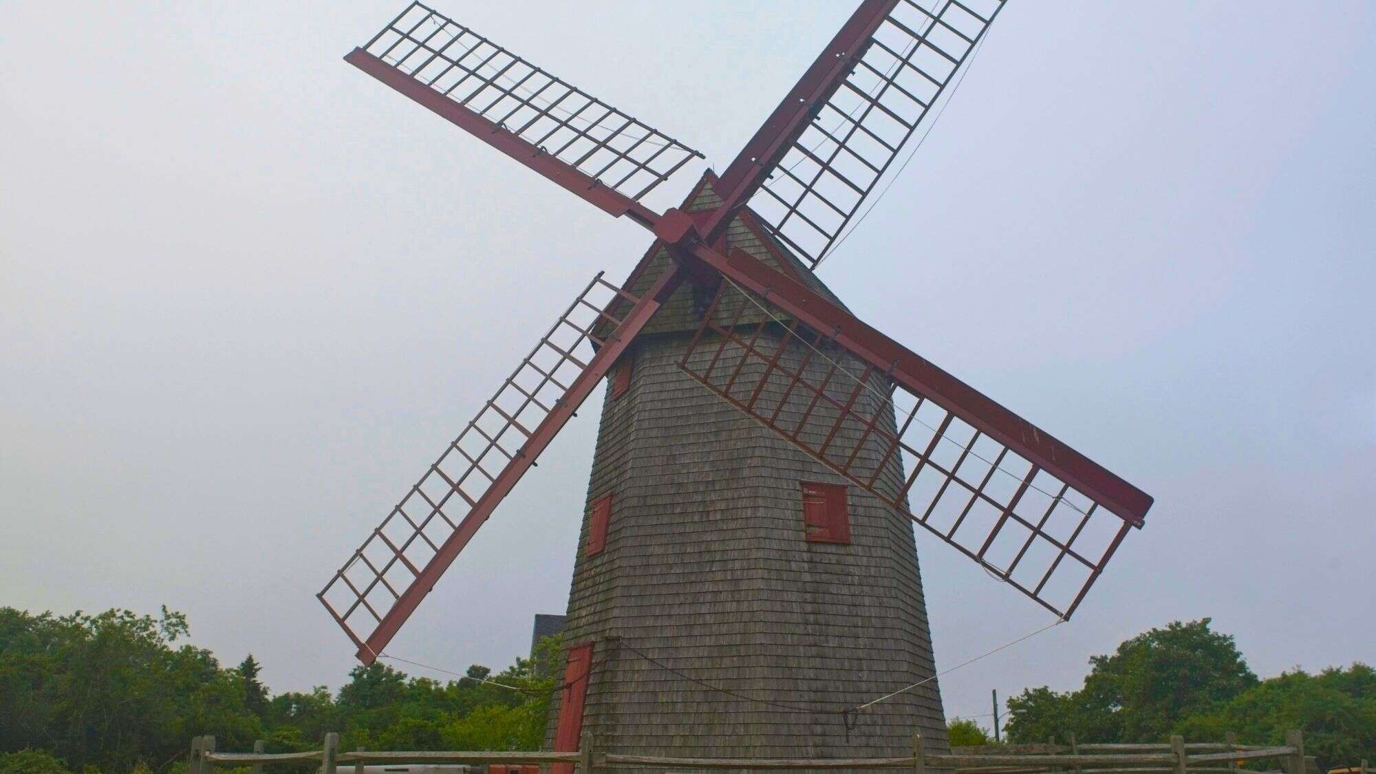 A rustic wooden windmill with red blades and shutters stands tall against a hazy sky, surrounded by greenery.