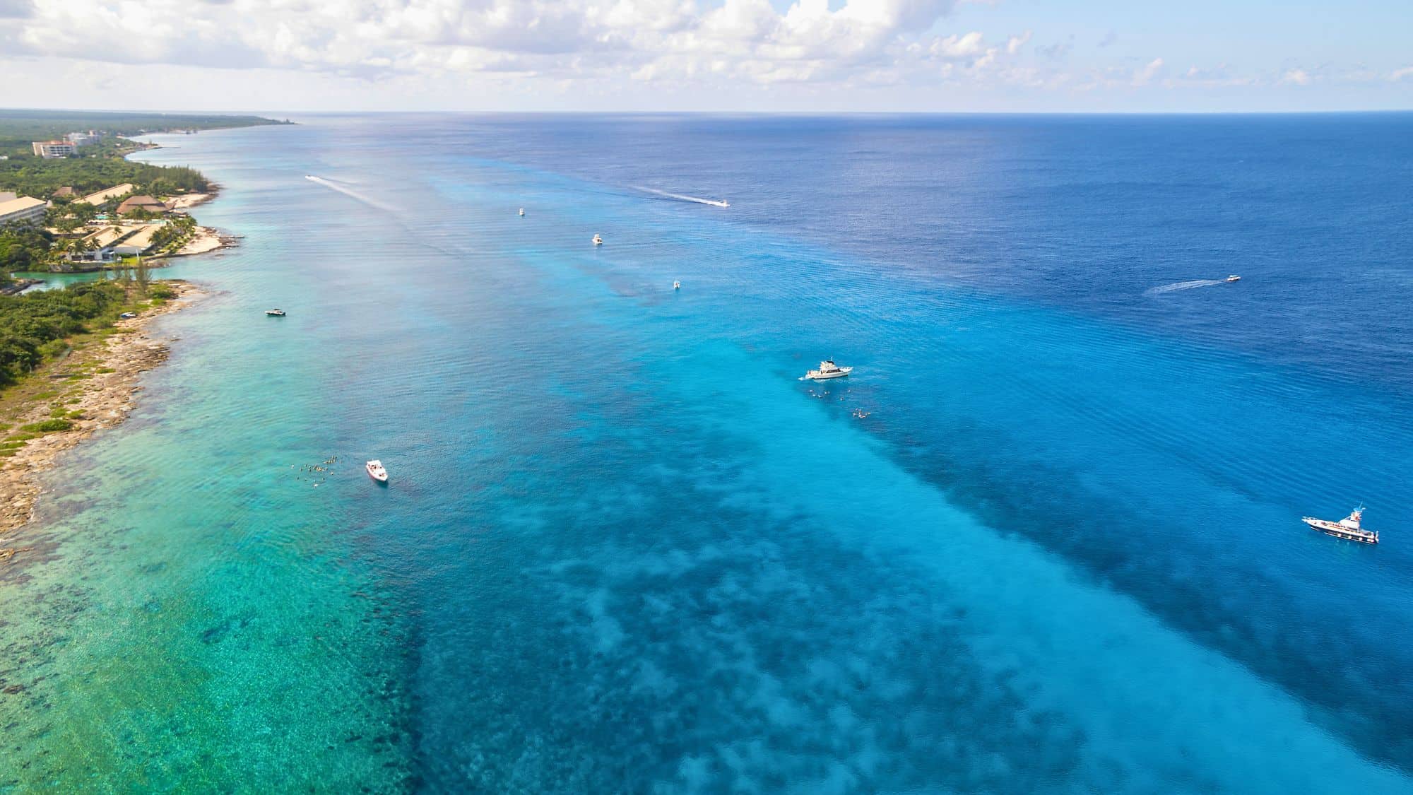 Boats dot the vivid blue waters along the shoreline of Cozumel, with resorts and greenery lining the coast.