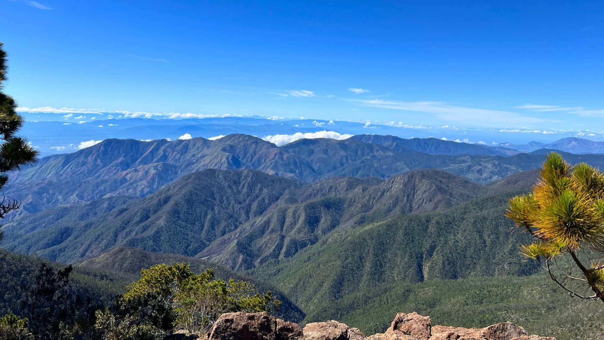 A sweeping view of layered mountain ridges covered in forest, fading into the horizon beneath a bright blue sky.