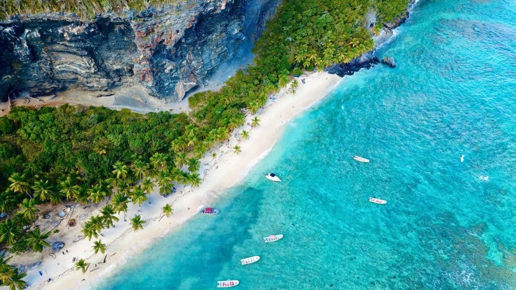 An aerial view of a white sand beach backed by palm trees and dramatic cliffs, with boats anchored in shallow turquoise water.