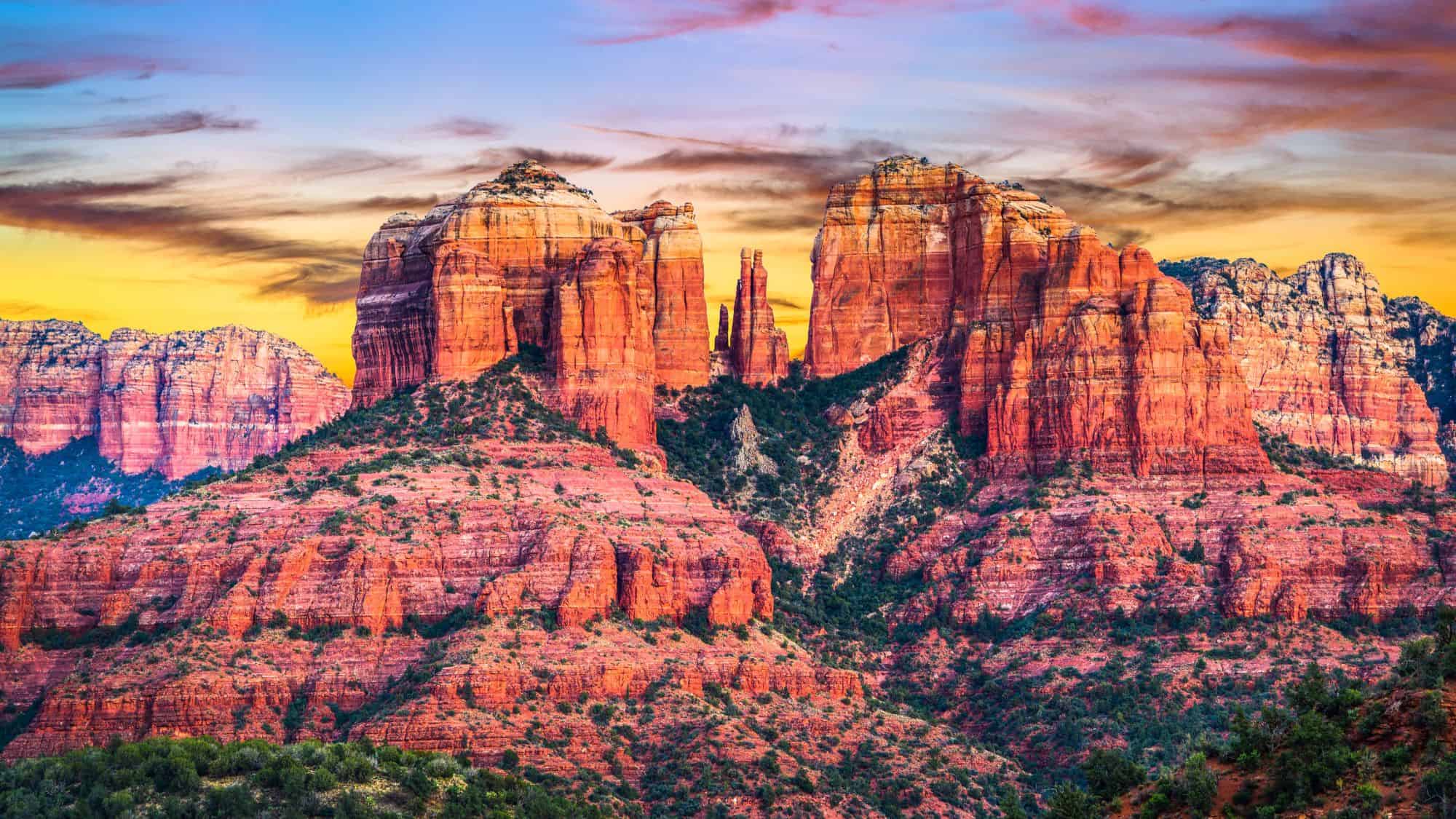 Towering red sandstone formations glow under a colorful sunset sky, with green vegetation scattered at their base.
