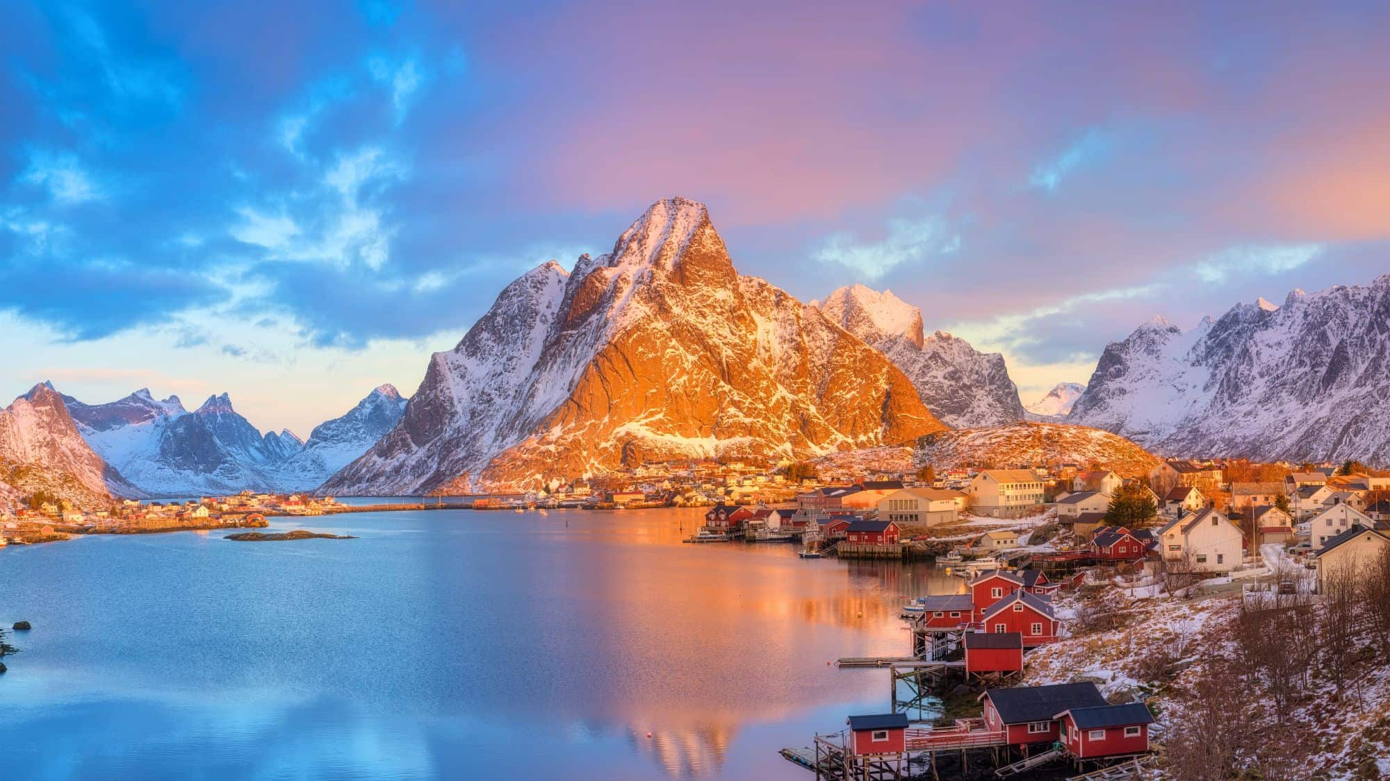 The village of Reine glows in the warm light of sunset, surrounded by snow-dusted peaks and calm waters.